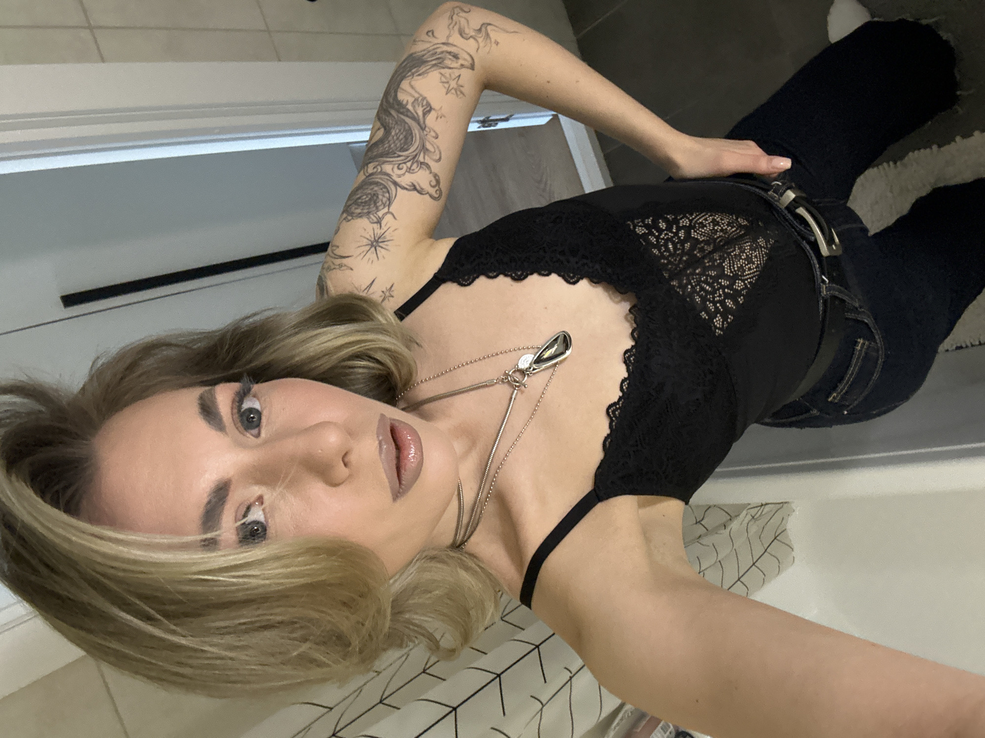 The image presents a female individual in an indoor setting within the city of Khimki, Russia. She is fair-skinned with blonde, shoulder-length hair and light-colored eyes, wearing light makeup including mascara, eyeshadow, and glossy neutral lipstick, complemented by a small piercing on her right nostril. Her attire consists of a black lace-trimmed camisole or bodysuit, dark blue jeans, and a black belt with a silver-colored buckle. She accessorizes with two silver-colored necklaces, one a choker and the other a longer chain featuring a teardrop-shaped pendant and a small round charm. Extensive black ink tattoo work covers her left arm, depicting a compass rose, stars, and complex swirling designs. The subject is captured in a selfie pose, holding the camera with her right hand (partially out of frame) and resting her left hand on her hip, gazing directly at the lens. The immediate environment includes a white shower curtain with a black geometric pattern on the left, a white fixture (possibly a bathtub or sink edge) below it, and white tiled walls and a doorframe to the right. The floor is dark grey tile, partially obscured by a white, shaggy rug.