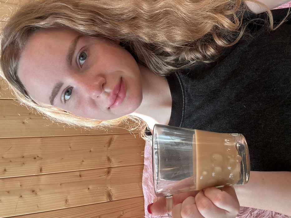 A young woman with light skin, blue-grey eyes, and long, wavy blonde hair is featured in a portrait-oriented photo, taken indoors in Vasilyevo, Russia. She is positioned on the right side of the frame, looking towards the camera with a gentle, calm expression. Her face is illuminated by natural daylight, highlighting her features and hair. She wears a dark, possibly black or charcoal gray, textured long-sleeved top. Her right hand, adorned with red painted fingernails, holds a clear glass mug containing a light brown, milky beverage, likely coffee, with some bubbles or ice visible. The background consists of light-colored wooden paneling, suggesting a rustic or cozy indoor setting like a wooden house or cabin. A glimpse of a light pink or purple fuzzy blanket is visible in the lower right portion of the image. There is no visible text.