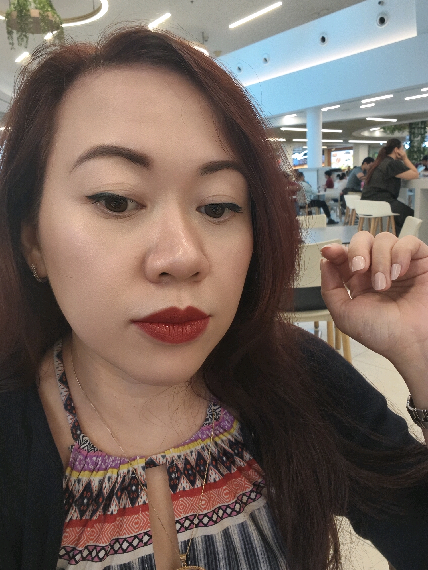 A medium shot captures a young Asian woman with long, dark auburn hair, wearing a black cardigan over a colorful, patterned top with a cutout at the neckline. She has on dramatic winged eyeliner and dark red lipstick, and is wearing a delicate gold necklace. Her right hand, with short, light-pink polished nails, is raised near her face, with her index finger extended as if to touch something. Her expression is neutral, and she is looking down and to the side. The background appears to be an indoor public space, possibly a food court or shopping mall, with bright overhead lighting and several blurred figures seated at tables in the distance. The overall lighting suggests an indoor environment with artificial illumination.