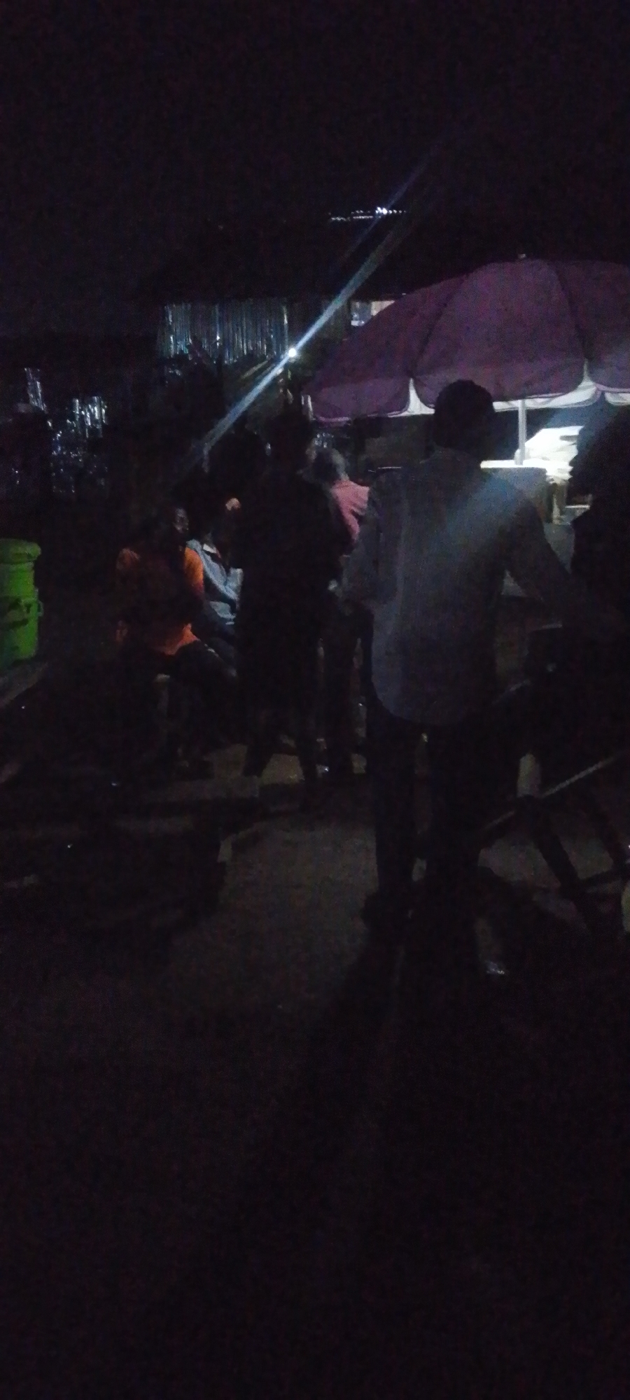The image depicts a nighttime outdoor scene in Gura Top, Nigeria, likely a market or street vendor area. In the foreground, a person wearing a light-colored shirt stands with their back to the camera, appearing to interact with someone to their right. Several other individuals are visible in the mid-ground, some seated and others standing, in various states of interaction. One person in the mid-left is wearing an orange shirt. There's a large purple umbrella or awning positioned in the upper right portion of the frame, suggesting a vendor setup. Various objects, including a green bucket on the far left, are present. The environment is dark, with artificial light sources creating some glare and illumination. The ground appears to be a rough, unpaved surface. The overall atmosphere suggests a lively, informal gathering.