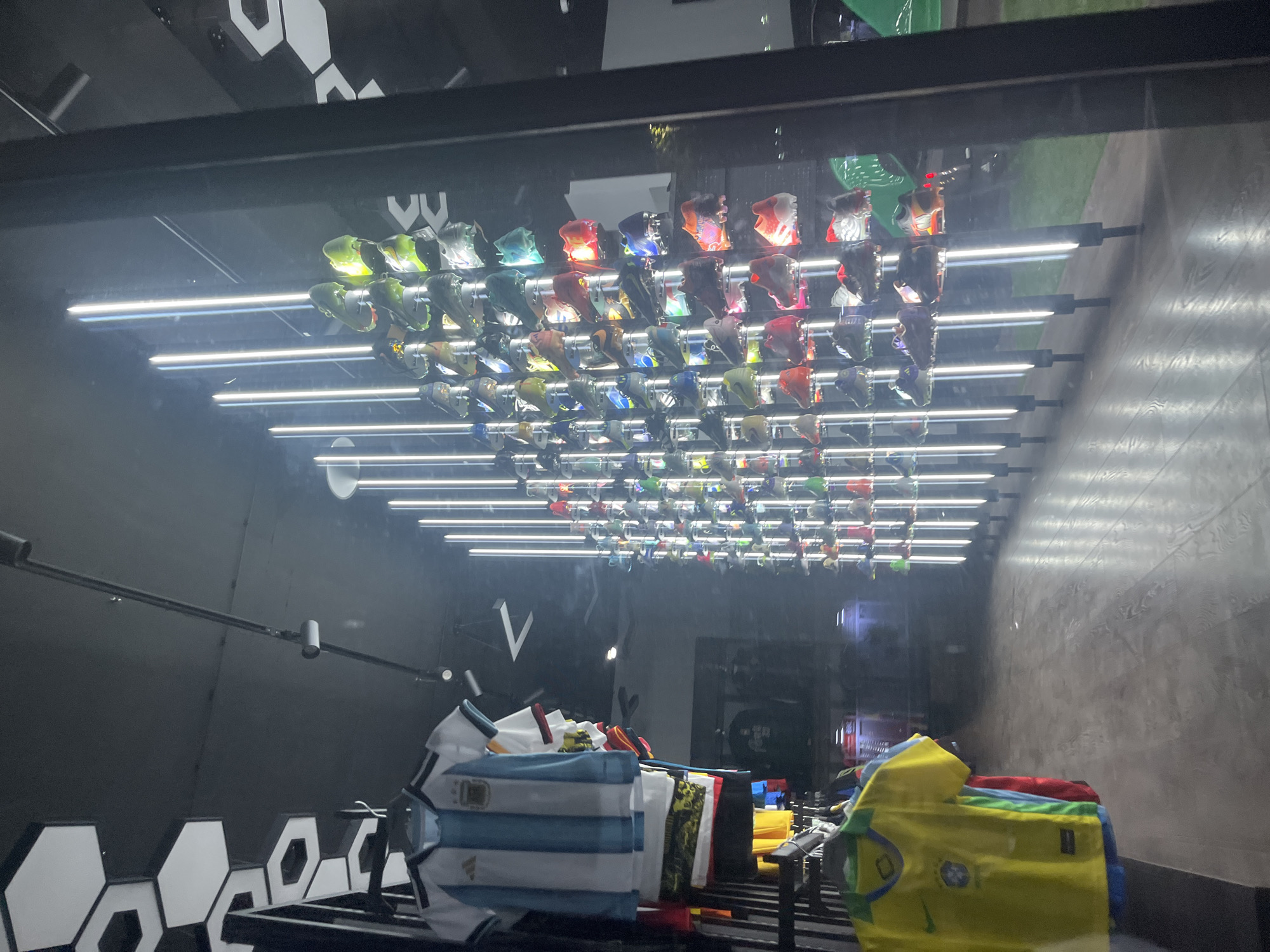 The image shows a display case in a store, likely in Yerevan, Armenia, given the user's specification. The case contains several soccer jerseys and a grid of soccer cleats hanging upside down from illuminated bars. The jerseys include recognizable teams like Argentina (white and blue stripes) and Brazil (yellow and green). The cleats vary in color. The background is a dark, patterned wall. The view is slightly obscured by reflections on the glass of the display case. The overall scene appears to be a retail environment showcasing soccer merchandise.