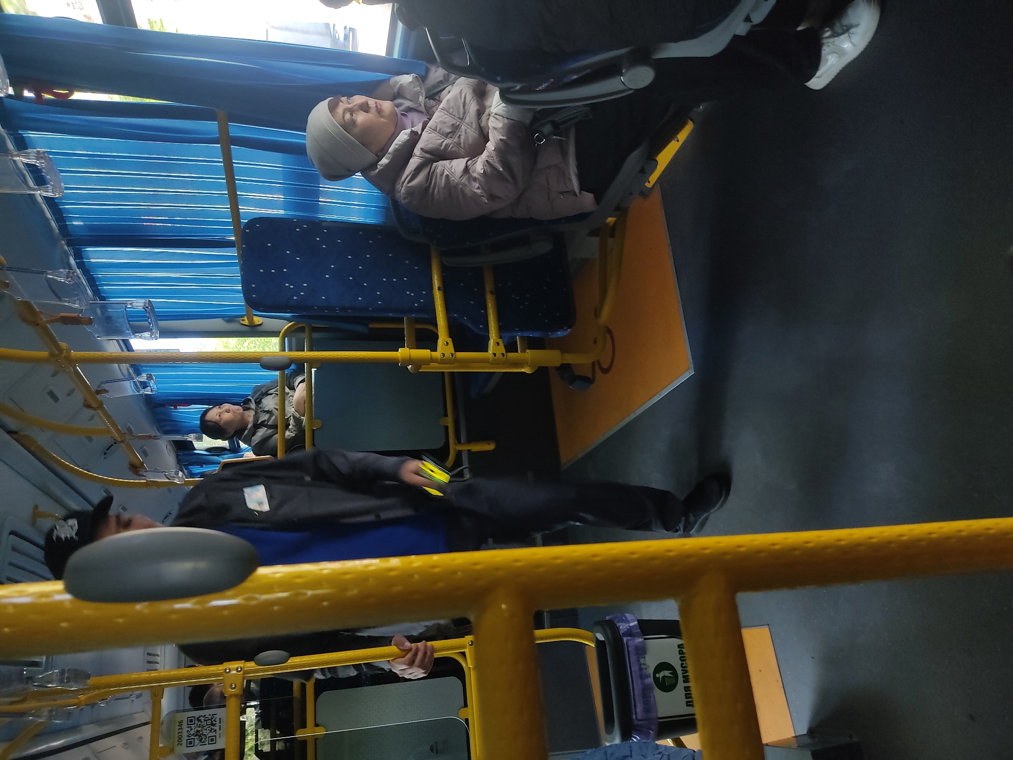 The image shows the interior of a bus. A woman in a grey hat and jacket is sitting on a blue seat with white dots, possibly sleeping. Further down the bus, another person is sitting. In the foreground, a man with a black cap is standing, holding what appears to be a phone. The bus has yellow support bars and blue curtains covering the windows. A sign with "для мусора" is visible. The location seems to be inside a public transportation vehicle.