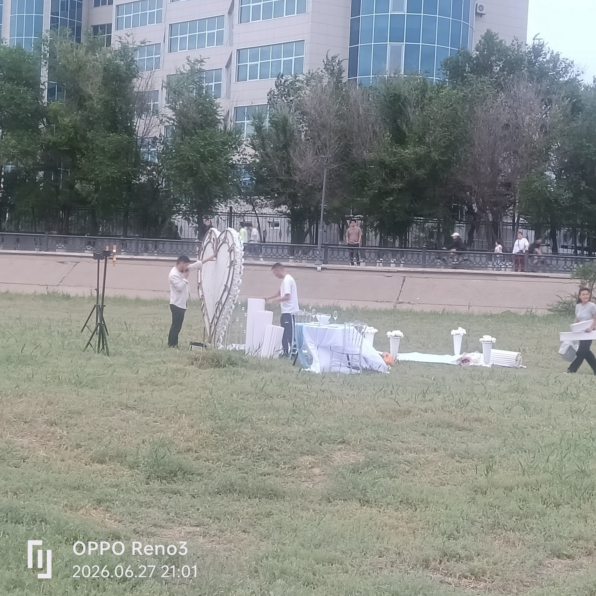 In Atyrau, Kazakhstan, an outdoor setup is being prepared in a grassy area with a modern building in the background. The scene appears to be for an event, possibly a celebration or romantic gesture, given the heart-shaped structure adorned with lights and flowers. Several people are present, engaged in arranging the decorations. One man in a white shirt and dark pants is adjusting the large heart structure, while another man in a white t-shirt and dark pants is working with white decorative pillars and a table set with white cloths, chairs, and glasses. A woman in a light-colored top and dark pants walks by carrying a white rectangular object. In the background, people can be seen walking and cycling along a paved path behind a decorative fence, with trees lining the area between the path and the building. The sky is overcast, suggesting a cloudy day. The time stamp in the lower left corner indicates the photo was taken on June 27, 2026, at 21:01.