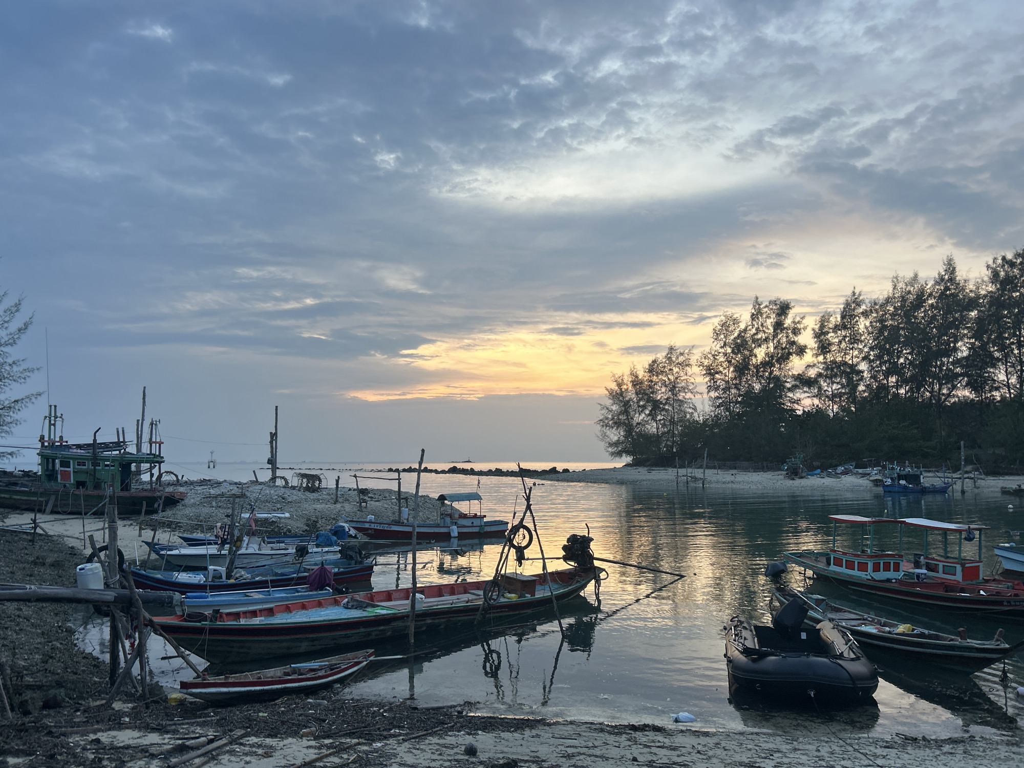 The image depicts a tranquil scene at ตำบลเกาะพะงัน, Thailand, likely near a coastline or harbor. Several boats of various sizes and types are moored or docked in the calm water. Some boats are traditional longtail boats, and one appears to be a small inflatable dinghy. The boats are reflected in the water's surface, creating a mirror-like effect that captures the soft light of the sky. The sky is overcast with a mix of grey and pastel shades, suggesting either sunrise or sunset. A line of trees is visible on the right side of the image, possibly indicating a nearby island or landmass. The foreground shows a sandy area, likely a beach, with natural debris scattered around. The overall mood of the image is peaceful and serene, capturing the essence of a quiet coastal setting.