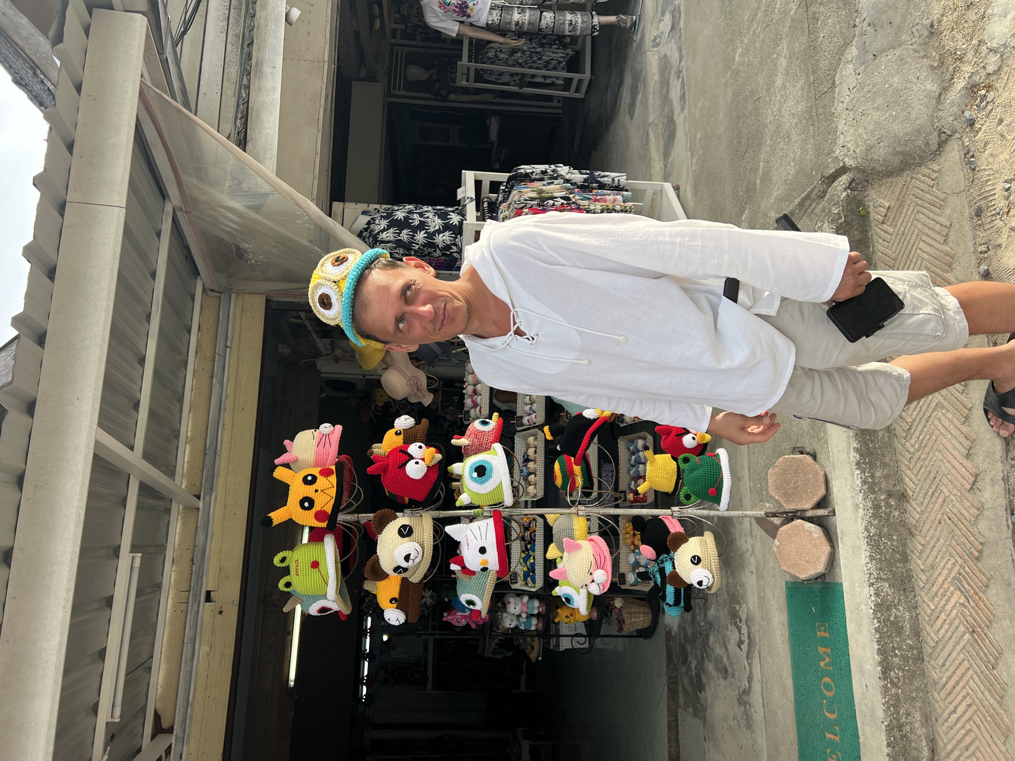 A light-skinned man in his late 20s or early 30s stands in front of a market stall in Baan Plai Laem, Thailand, wearing a white hooded tunic and light-colored shorts. He has a playful expression and is wearing a novelty hat featuring two large, bulging eyes. The stall behind him displays a variety of colorful, crocheted hats shaped like cartoon characters and animals, including a frog, Pikachu, a panda, and an owl. The man is holding a black rectangular object, possibly a phone or wallet, in his right hand. The background shows a street scene with more market stalls and clothing racks. The lighting suggests it is daytime, possibly late afternoon due to the long shadows. The man is the primary subject and is looking towards the camera.