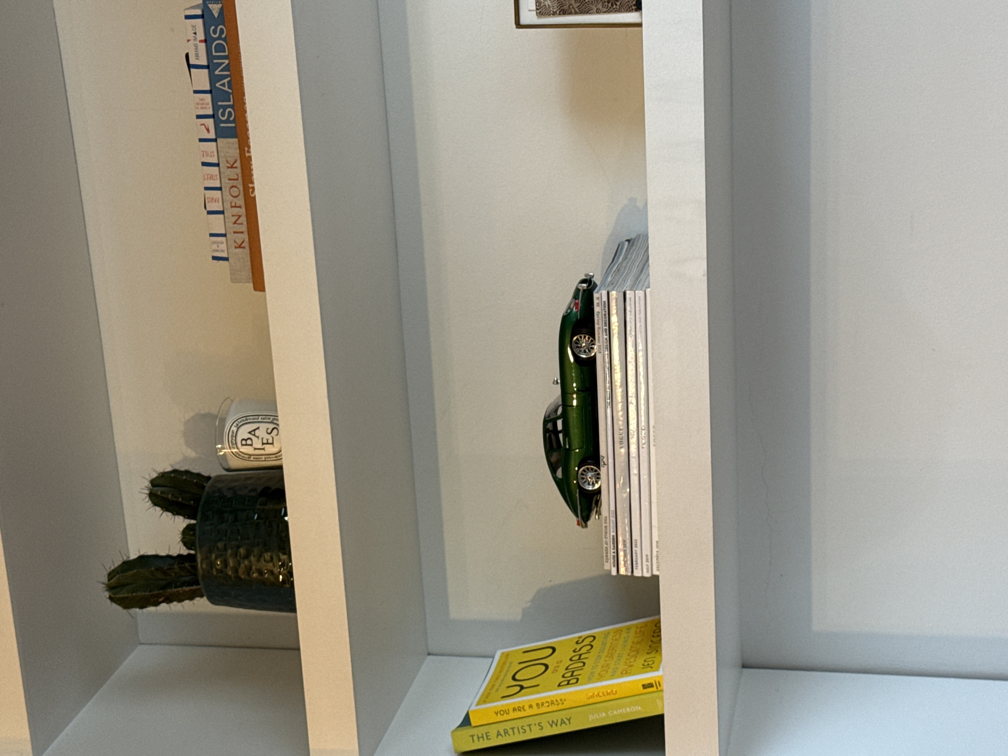 An interior scene depicts a white, multi-tiered shelving unit against a plain white wall.
On the top shelf, positioned to the left, a dark green textured pot contains three distinct green cacti. To its right, a white cylindrical Diptyque candle with a black 'BAIES' label is present. Further right, a horizontal stack of five books includes visible titles such as 'KINFLK' and 'ISLANDS'.
The middle shelf, from left, displays two yellow upright books titled 'THE ARTIST'S WAY' and 'YOU ARE A BADASS'. Adjacent to these is a horizontal stack of approximately eight magazines or thin periodicals, topped by a dark green model sports car, resembling a Jaguar E-Type, oriented to the right. A small portion of a gold-framed item is visible on the far right of this shelf.
The bottom shelf is empty and white. No individuals are present. The environment suggests a domestic or commercial interior space in London, United Kingdom.