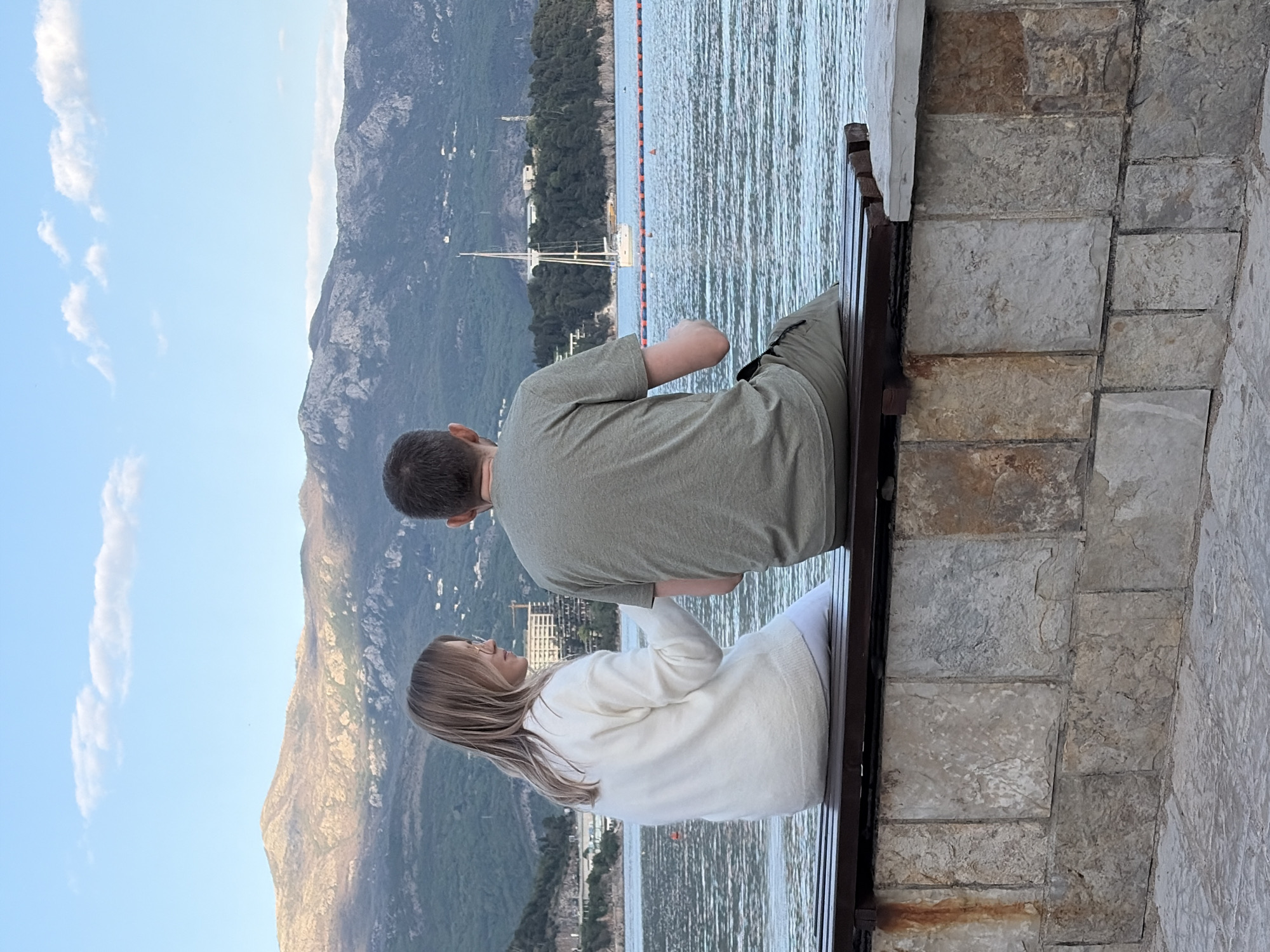 Two people are sitting on a bench. A woman with long, blonde hair is wearing a white sweater. A man beside her is wearing a green t-shirt. They are on a bench constructed of stone, facing the sea, with mountains in the background. A sailboat sits on the water in the background, behind an orange buoy line. To the right of the bench is a wall constructed of stone. They appear to be in Budva, Montenegro.