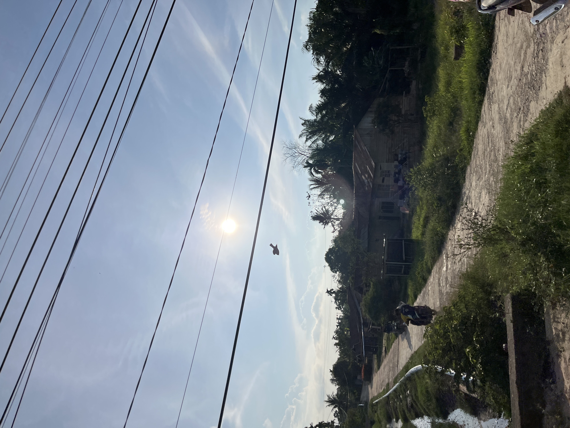 The image depicts a rural scene in Alurcucur, Indonesia, under a bright, sunny sky with scattered clouds. Power lines are visible crisscrossing the upper portion of the frame. In the sky, a small object, possibly a kite or a distant drone, is flying. The landscape features a dirt road leading into a village, with several modest houses with tin roofs. Lush green vegetation, including palm trees, borders the road and surrounds the dwellings. On the road, a person is riding a motorbike away from the viewer. To the left of the road, a narrow body of water, likely a canal or irrigation ditch, is present. The overall impression is one of everyday life in a tropical village.