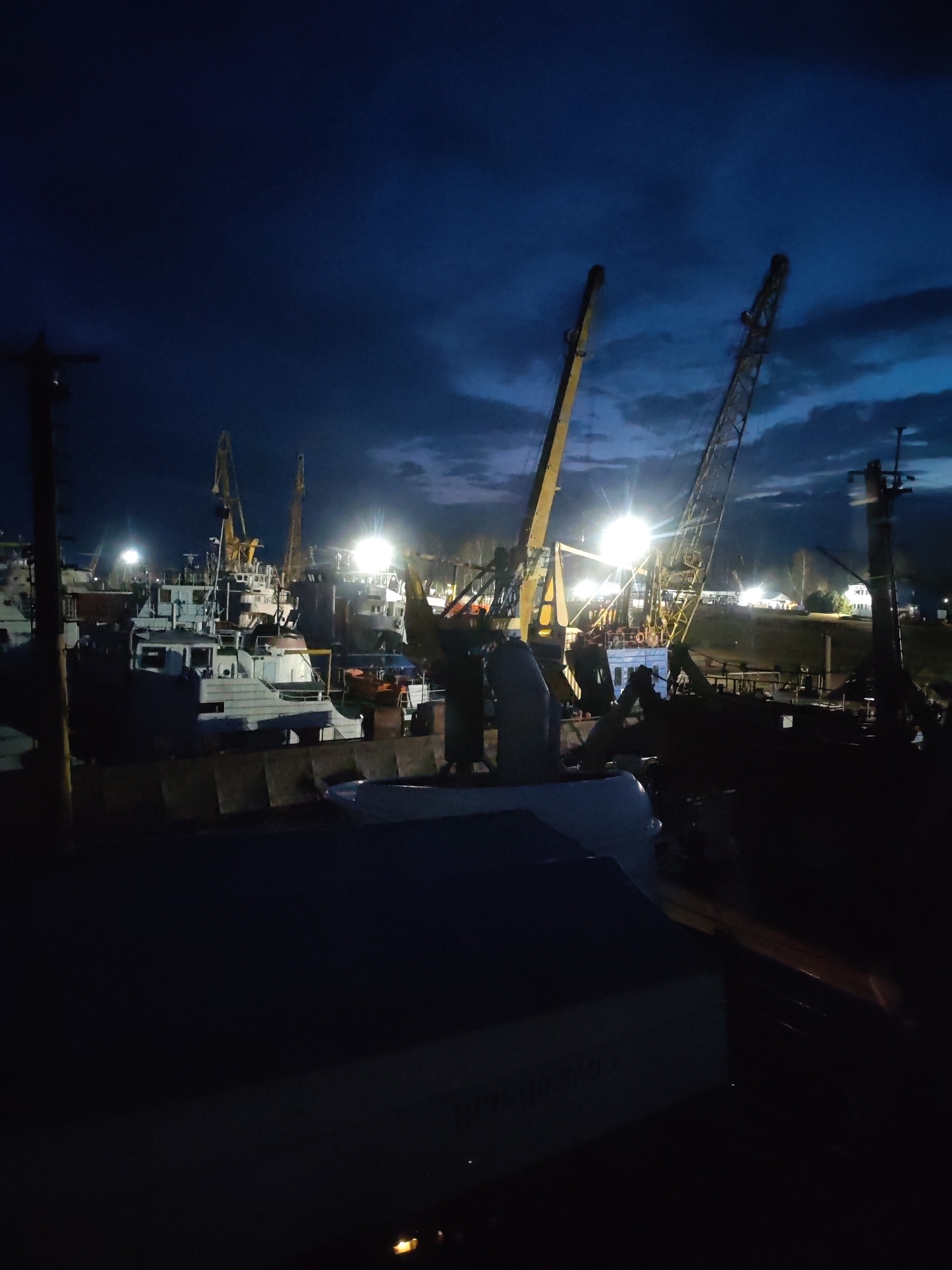 The photograph depicts a dark, nighttime scene at a dock or shipyard in Rybinsk, Russia. Numerous boats and vessels are visible, some illuminated by bright lights, with cranes towering above them. The sky is a deep blue with scattered clouds, and some lights are visible in the distance on what appears to be elevated ground. The foreground is dark, suggesting the photographer is on another boat or structure.