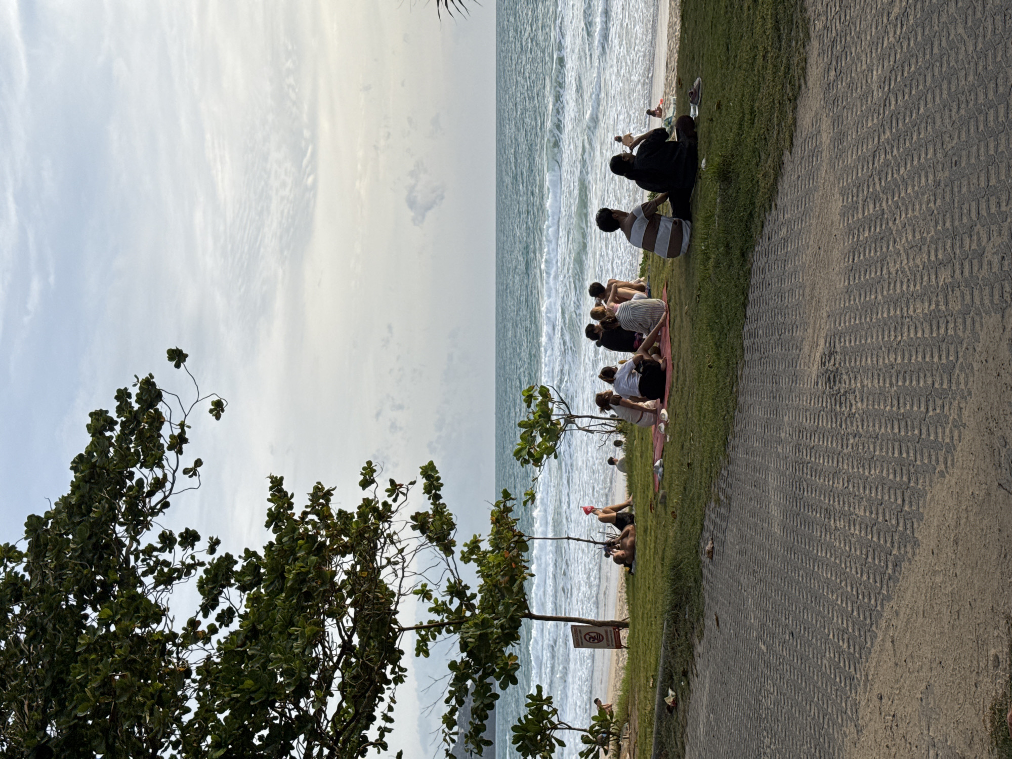 An outdoor, eye-level, medium shot captures a serene beach scene in Choeng Thale, Thailand, during what appears to be late afternoon or early evening. The foreground is dominated by a textured gray paved pathway, leading towards a grassy area where several individuals are gathered.
On the grass, approximately eight people are seen sitting, mostly facing the ocean. Some are arranged in small groups, while others are more spread out. One person in the middle ground is lying down on the grass, wearing a red head covering. Another individual, also in the middle ground, is wearing a white top and dark bottoms, appearing to be engaged in conversation with another person. In the foreground, a group of five people are seated on a red mat, facing away from the camera. One person in this group is wearing a striped shirt. Further to the right, two individuals are seated, one wearing glasses and a dark top, the other in a striped shirt.
Beyond the grassy area, a sandy beach stretches towards the turquoise ocean, where moderate waves are breaking. A few people are visible on the beach, further in the distance. A small, dark-colored signpost with red markings is situated near the edge of the grass.
The background is filled with the expansive ocean and a sky with scattered clouds, tinged with the soft light of a setting sun. Large trees with dense green foliage frame the left side of the image, with their branches extending over the scene. The overall atmosphere is one of relaxation and leisure, with people enjoying the natural beauty of the coastal environment.