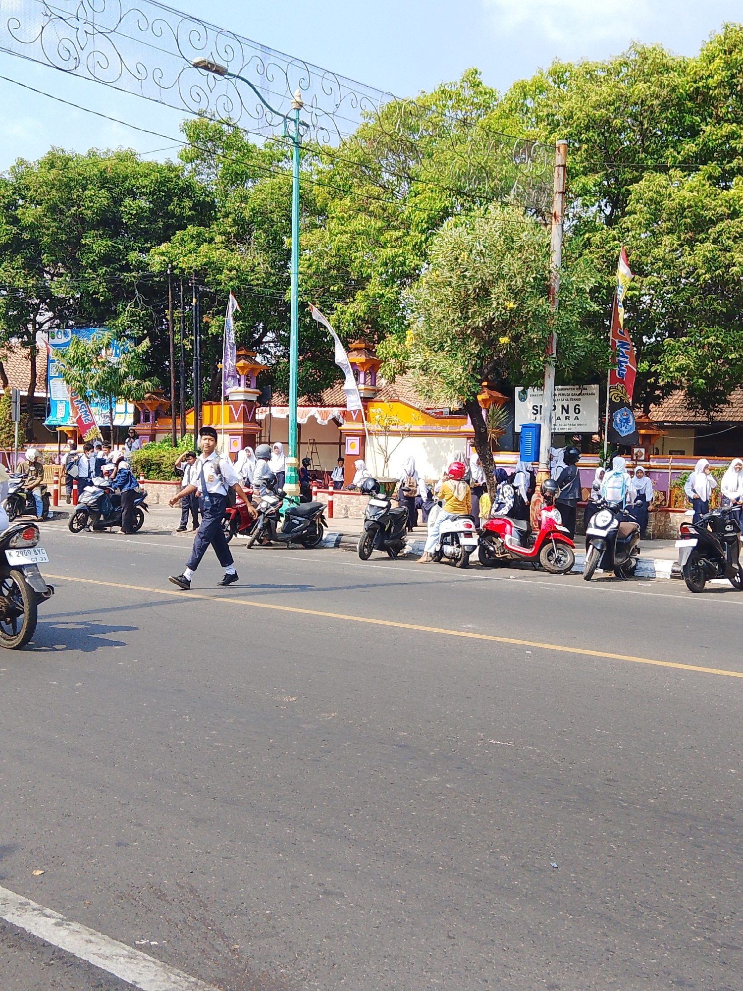 The image captures a street scene in Jepara, Indonesia, likely near a school. In the foreground, a young male student in a white shirt, blue pants, and a white backpack is walking across the street. Several motorcycles are parked on the side of the road. In the background, more students, mostly wearing white uniforms and headscarves, are gathered near buildings that appear to be part of a school campus, indicated by signage that reads "SMP N 6 JEPARA". The buildings have traditional Indonesian architectural elements, with orange and yellow accents. Lush green trees line the street and campus. The sky is clear and blue, suggesting a sunny day. The overall scene depicts a typical daily activity of students in Indonesia.