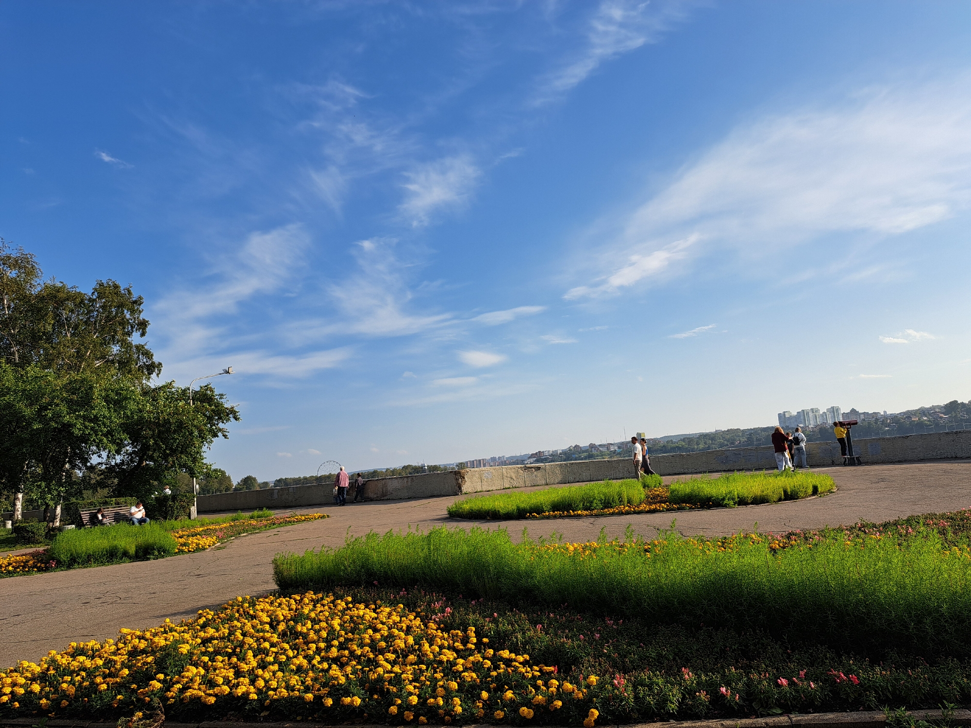 The image depicts a scenic view of a park in Irkutsk, Russia, likely overlooking the Angara River. In the foreground, a paved walkway winds through vibrant flower beds bursting with yellow marigolds. Tall, lush green grass borders sections of the walkway, creating a layered effect.
Several people are visible in the park. On the left, a person is seated on a park bench beneath a leafy tree. Further along the walkway, a group of individuals are strolling and observing the view. One person is standing at a public telescope, appearing to be taking in the panoramic cityscape in the distance. The cityscape features modern buildings and hints of greenery, suggesting it is a well-established urban area.
The sky above is a bright, clear blue, adorned with wispy white clouds that add depth and texture to the scene. A tall lamppost stands near the trees on the left, and in the far distance, a Ferris wheel is partially visible, hinting at an amusement park or entertainment area nearby. The overall atmosphere is peaceful and invites leisurely exploration of the park's natural beauty and city vistas.