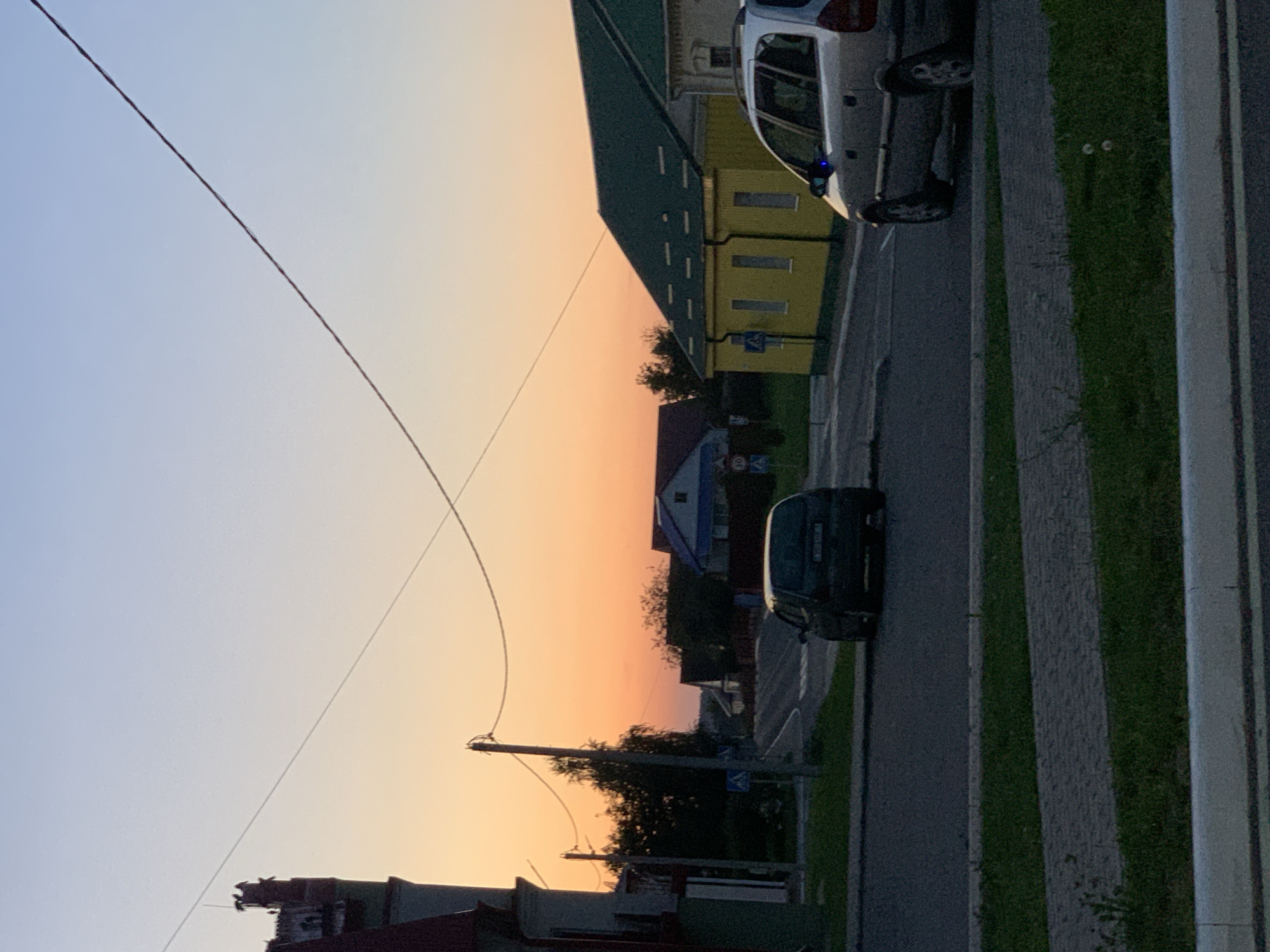 The image depicts a street scene in Kapyĺ, Belarus, during what appears to be dusk or early evening. The sky exhibits a gradient of colors, transitioning from a pale blue at the top to a warm orange and yellow near the horizon. A single utility pole with wires stretches across the upper portion of the frame.
In the foreground, a paved road runs horizontally. To the left, a dark-colored hatchback car is parked on the side of the road. Further back, a larger, silver station wagon is visible on the right side of the road, facing away from the viewer.
Several buildings are present in the background. A distinctive yellow building with a green roof and white-trimmed windows is situated on the right side of the road. To its left, a residential house with a blue roof is partially visible. A dark green tree line separates the road from the cluster of houses in the distance. A tall, narrow building with a dark roof and red accents, possibly a tower or an old structure, is visible on the far left side of the image, partially obscuring a light-colored building behind it. Road signs, including a pedestrian crossing sign, are visible along the street. The ground on either side of the road is covered with grass and paved walkways.