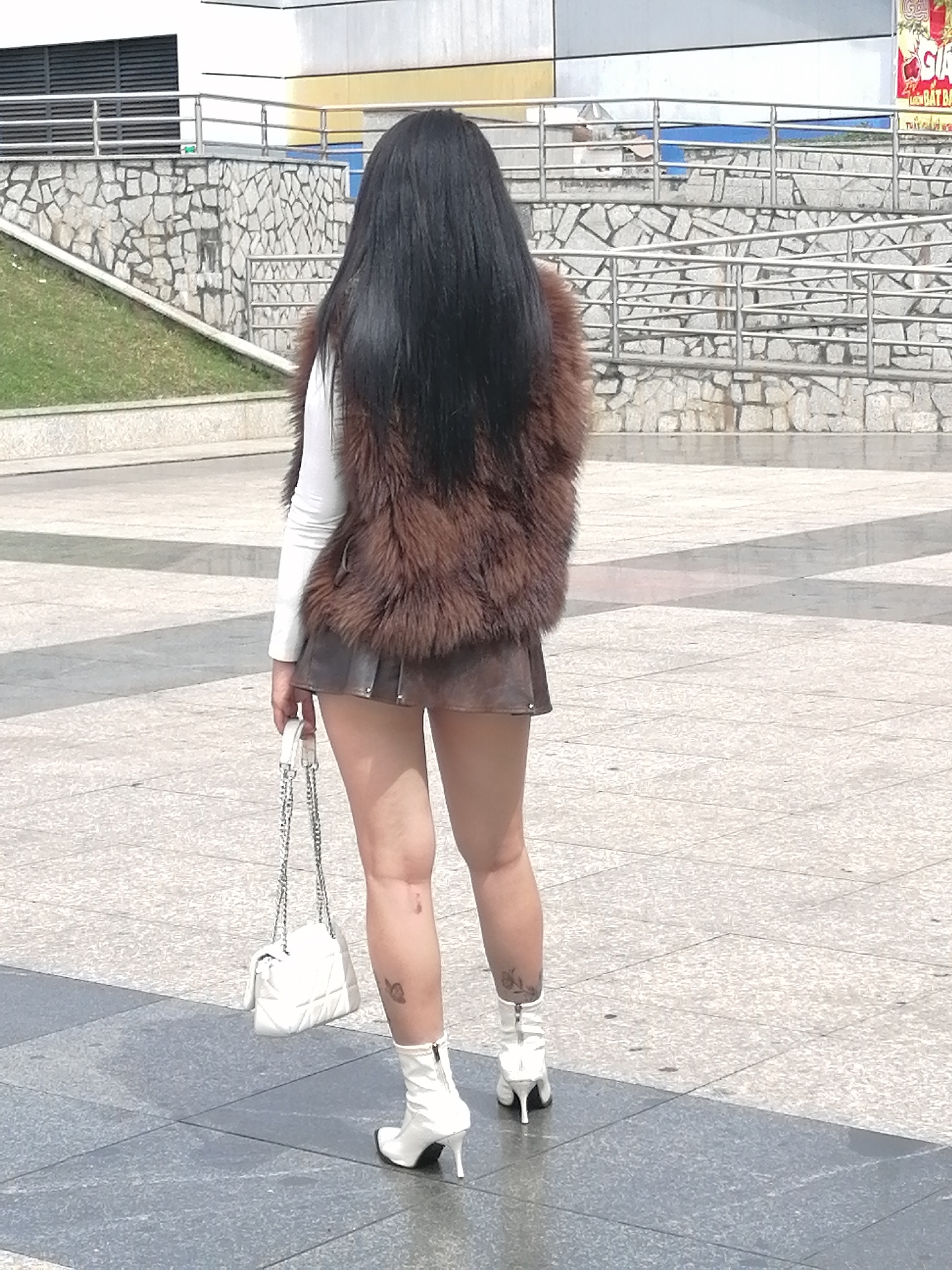 A person stands outdoors, facing away from the camera, in what appears to be a public plaza in Da Lat, Vietnam. The person has long, dark hair and is wearing a brown fur vest over a white long-sleeved shirt. Below the vest, they are wearing a short, dark skirt. They are holding a white quilted handbag with a silver chain strap in their left hand. The person is also wearing white heeled ankle boots, and has visible tattoos on their legs. The ground is paved with large, light-colored tiles, with some darker, rectangular tiles interspersed. In the background, there is a stone wall with a metal railing, and a grassy slope leading up to a building with a yellow and white facade. The lighting suggests it is daytime.
