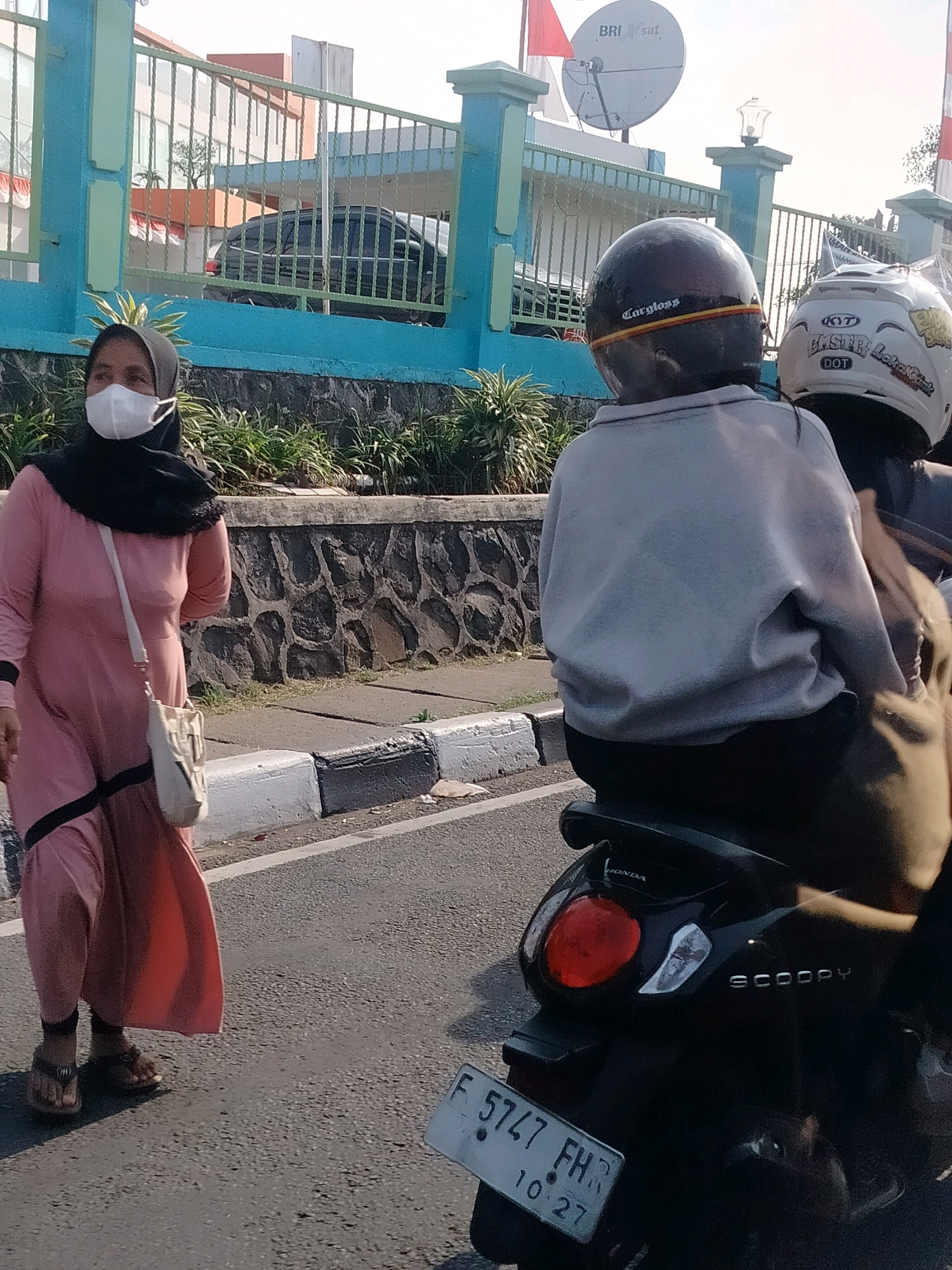 In Cisarua, Indonesia, on a sunlit day, a woman wearing a pink dress, black hijab, and a white face mask walks along a paved road. She carries a light-colored sling bag. Behind her, two individuals on a black scooter, one wearing a grey sweatshirt and the other a white helmet, are visible. The scooter has a license plate reading "F 5747 FH" and "10 27". The background features a blue and green fence, a stone wall, and a building with orange accents. The scene appears to be a typical street view in a residential or commercial area.