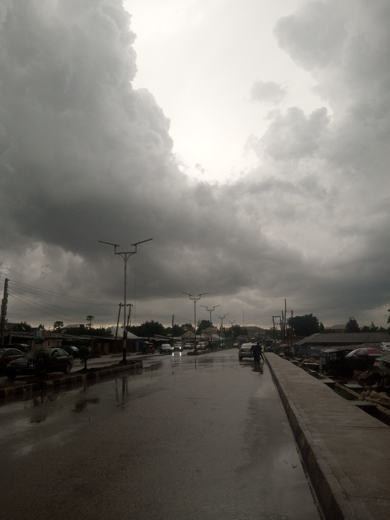 This media file captures a street scene under heavy, dark clouds, suggesting impending or ongoing rain. The sky is filled with dramatic, textured clouds that dominate the upper portion of the frame. The wet road reflects the overcast sky and the lights of vehicles.
The scene features a paved road with several cars driving on it. On either side of the road, there are buildings that appear to be shops or residences. Some have awnings or canopies, and there are indications of market stalls or informal vendors on the right side, with umbrellas visible. Lampposts are spaced along the road, one particularly prominent one near the center. A few trees are scattered throughout the scene.
The presence of wet surfaces and the dark, cloudy sky strongly indicate rainy weather, possibly just after a downpour or during a light rain. The overall mood is somewhat somber and subdued due to the weather. The time of day is difficult to determine precisely but appears to be late afternoon or early evening, given the dim lighting and the atmospheric conditions. There is no visible text in the image.