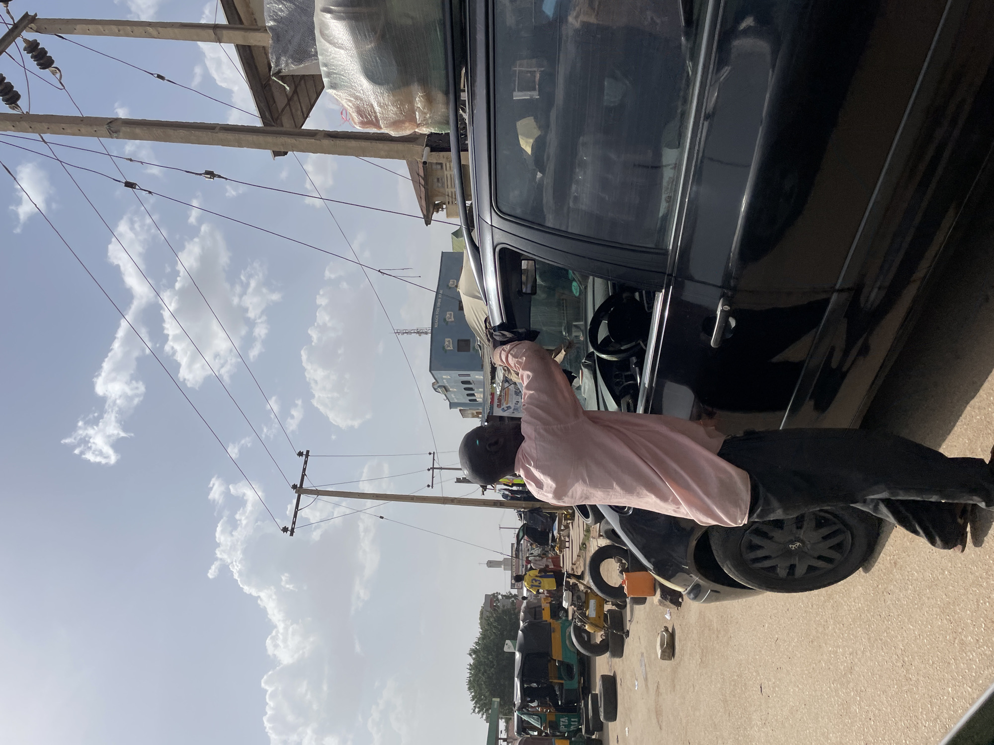 This daytime image captures a bustling street scene in Jos, Nigeria, under a bright sky with scattered white clouds. The central subject is a man with light brown skin and a bald head, dressed in a light pink long-sleeved shirt and dark trousers. He is leaning into the open driver's side window of a dark-colored car, which has items, possibly luggage or goods, secured to its roof.
The paved street is active, with several auto-rickshaws (commonly known as Keke Napep in Nigeria) visible in the background, along with other people and buildings. Wooden utility poles with numerous wires run across the upper part of the frame. One distinct light blue building in the background features a white signboard with visible text that reads "BALAKA HOUSE" and "NO. 0005 TEL:". On one of the auto-rickshaws, "JBPZA" and "2064" are partially legible. A small plastic container rests on the ground near the car's rear wheel.
The activity appears to be an everyday interaction, perhaps a transaction, a conversation, or a preparation for travel, consistent with a busy urban or semi-urban street environment. The natural light suggests it is mid-morning or afternoon.