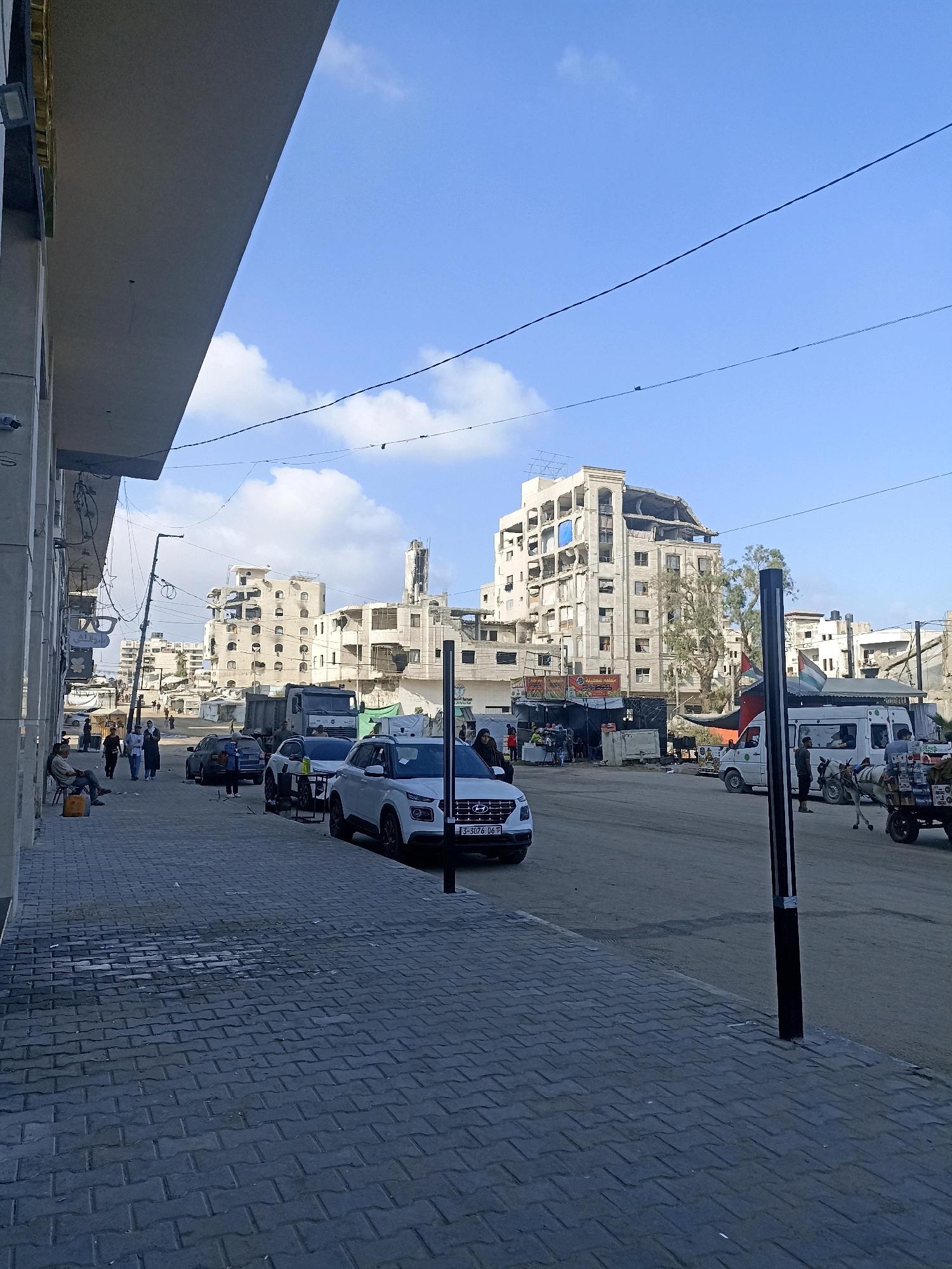 A street scene in the city of Gaza, Palestinian Territories, shows a paved sidewalk on the left with a man sitting on a low stool and a few people walking in the background. The sidewalk leads to a street with parked cars, including a white Hyundai SUV, a white van, and a dump truck. Several buildings line the street, some appearing damaged or under construction. In the distance, more multi-story buildings are visible, some with significant structural damage. The sky is blue with scattered clouds, and power lines run across the scene. A horse-drawn cart with goods is moving on the right side of the street.