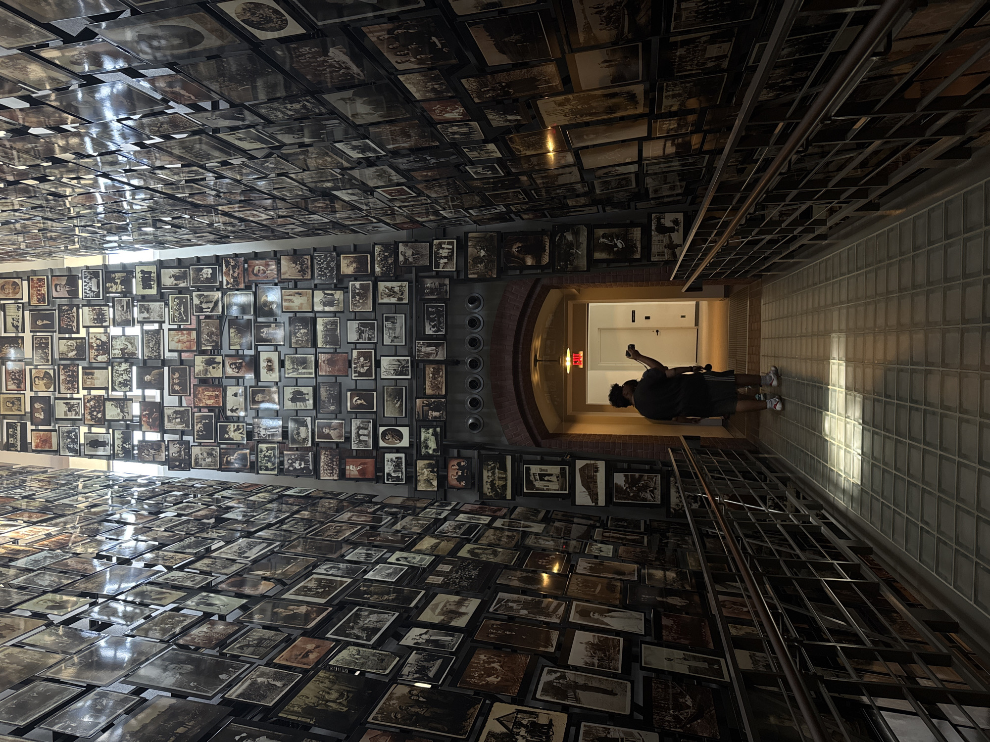 The image depicts a museum exhibit with a vast collection of black and white photographs displayed on walls and an expansive floor installation. The photographs appear to be historical, showing people in various settings, possibly from the early to mid-20th century. The walls are covered with framed images, creating a mosaic effect, and the floor is covered with rows of unframed photographs, laid out on metal racks.
In the background, a person is visible walking away from the camera, towards a brightly lit hallway. This person is dressed in casual attire and appears to be a visitor to the exhibit. The lighting in the scene suggests it is daytime, with natural light filtering in from an unseen source, creating strong shadows on the floor and illuminating the glass block wall on the right. The overall atmosphere is somber and contemplative, fitting for a historical exhibition. There is no visible text in the image. The location is identified as Washington, United States of America.