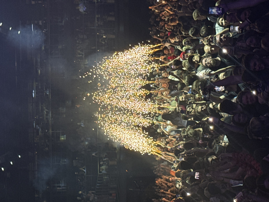 The image depicts a large indoor concert or live event within the VTB Arena – Dynamo Central Stadium in Moscow, Russia. A dense crowd of individuals occupies the foreground and midground, with additional spectators visible in multiple dark, tiered seating sections extending into the upper background. Approximately seven distinct plumes of golden pyrotechnic sparks are actively erupting upwards from handheld devices within the main standing crowd, illuminating the immediate area and producing a visible haze. Numerous individuals in the foreground are holding mobile phones aloft, many with activated screens or camera flashes, apparently recording or photographing the event. People in the crowd are generally facing forward, with many arms raised. The overall environment is dark, with minimal ambient lighting beyond the pyrotechnic displays and individual phone lights. Dark structural elements and overhead rigging are discernible in the upper sections of the venue.
