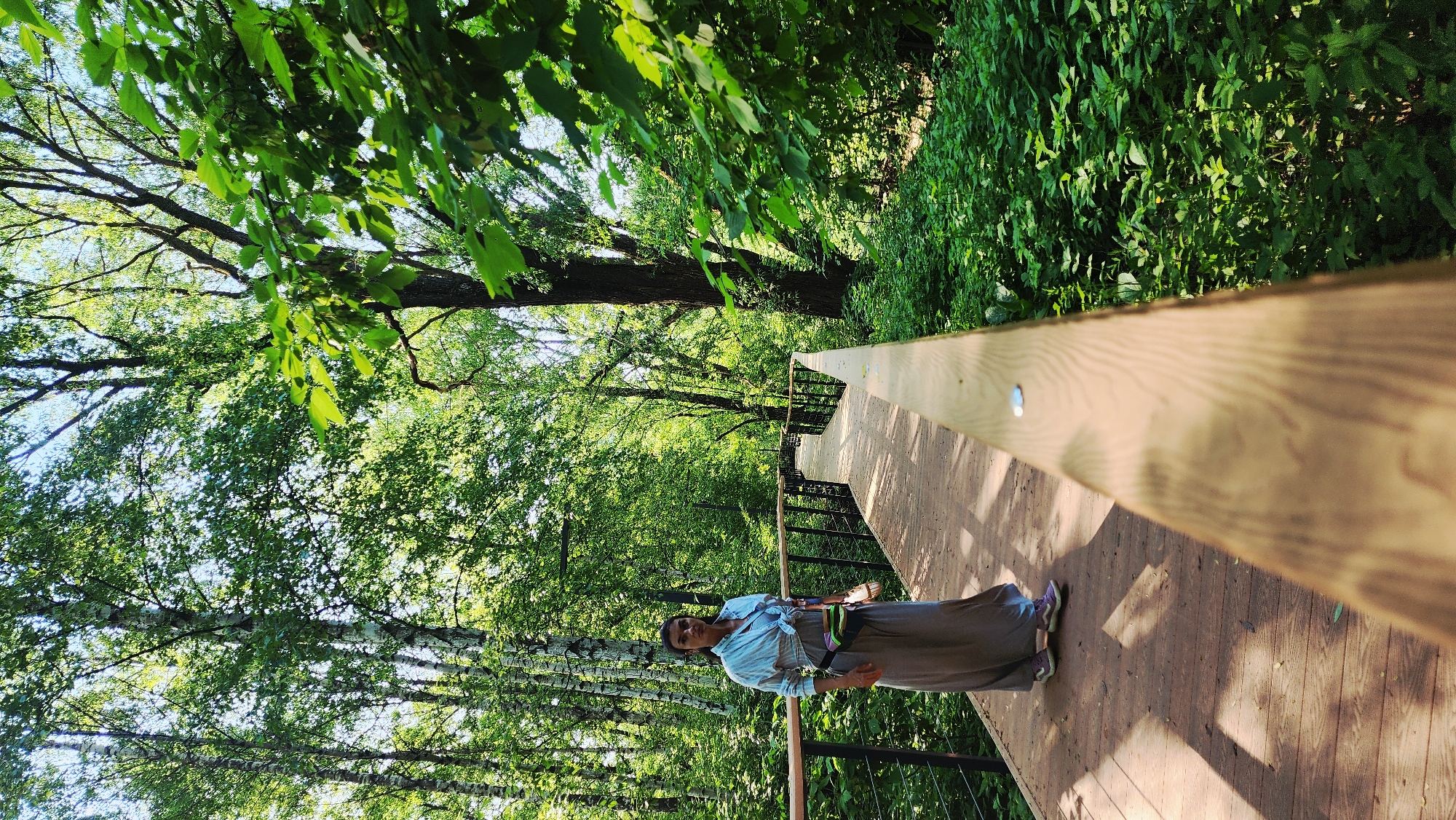 A woman stands on a wooden walkway in a wooded area, with trees and lush greenery surrounding her. The walkway is elevated and has wooden railings on either side. The woman is wearing a long grey skirt, a light blue shirt tied at the waist, and purple shoes. She has a dark fanny pack on her waist and a water bottle in her hand.
Based on the environment, this image is likely from the "Trail Among the Trees" in VDNKh Park, Moscow, Russia.