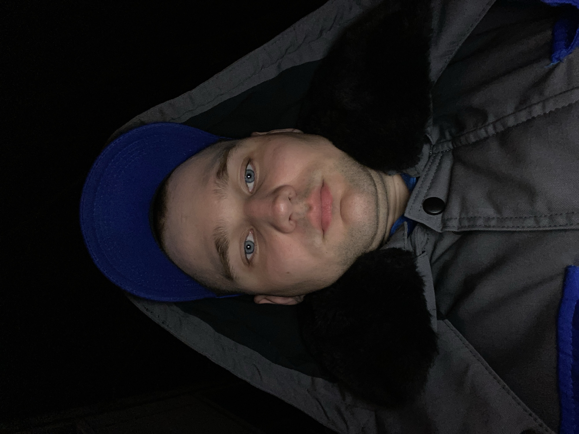 The image displays a frontal, close-up portrait of an adult male. The subject has fair skin, blue eyes, and light stubble on his chin and cheeks. He is wearing a blue baseball cap and a gray hooded jacket with a black, possibly faux-fur, collar. The jacket's hood is pulled up, partially obscuring the sides of his head. A dark, circular button is visible on the right lapel area of the jacket, and a segment of a blue undergarment is visible below the jacket opening on the lower right. The background is uniformly black and featureless, providing no environmental context. The specific location within Chaikovsky, Russia, cannot be determined from the visual information presented in the image.