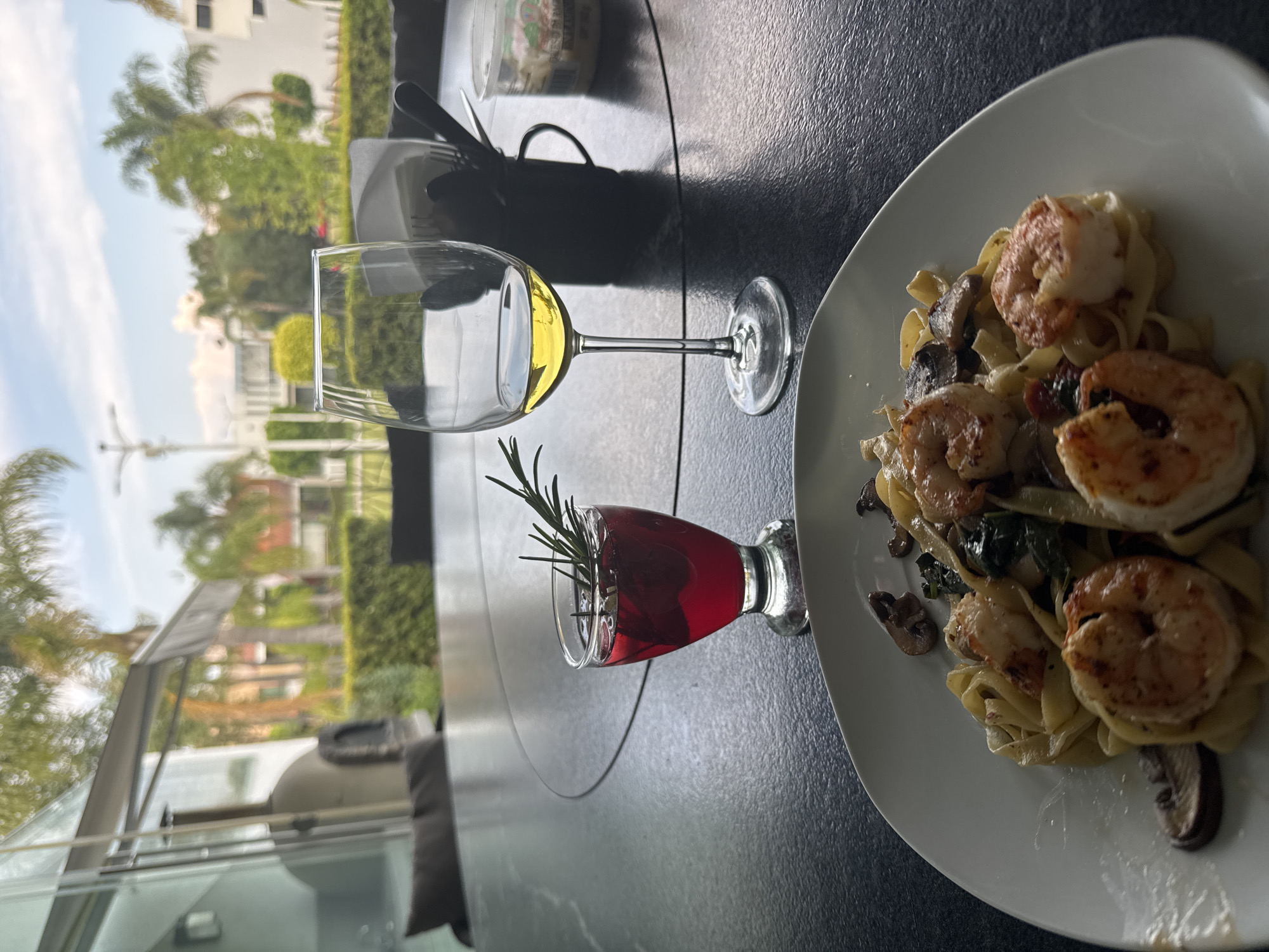 The image depicts a table setting outdoors in San Bernardino Tlaxcalancingo, Mexico, based on the identifiable architectural styles and local flora visible in the background. A plate of pasta with shrimp and mushrooms is prominently displayed on a white plate. To the left, there are two glasses: one containing a yellow liquid, possibly white wine, and the other containing a red liquid, perhaps a cocktail or red wine, garnished with a sprig of rosemary. A set of silverware in a napkin and a glass container are also present on the dark-colored table. The background features vegetation and buildings, typical of the urban landscape in the region. There are no people visible in the image.
