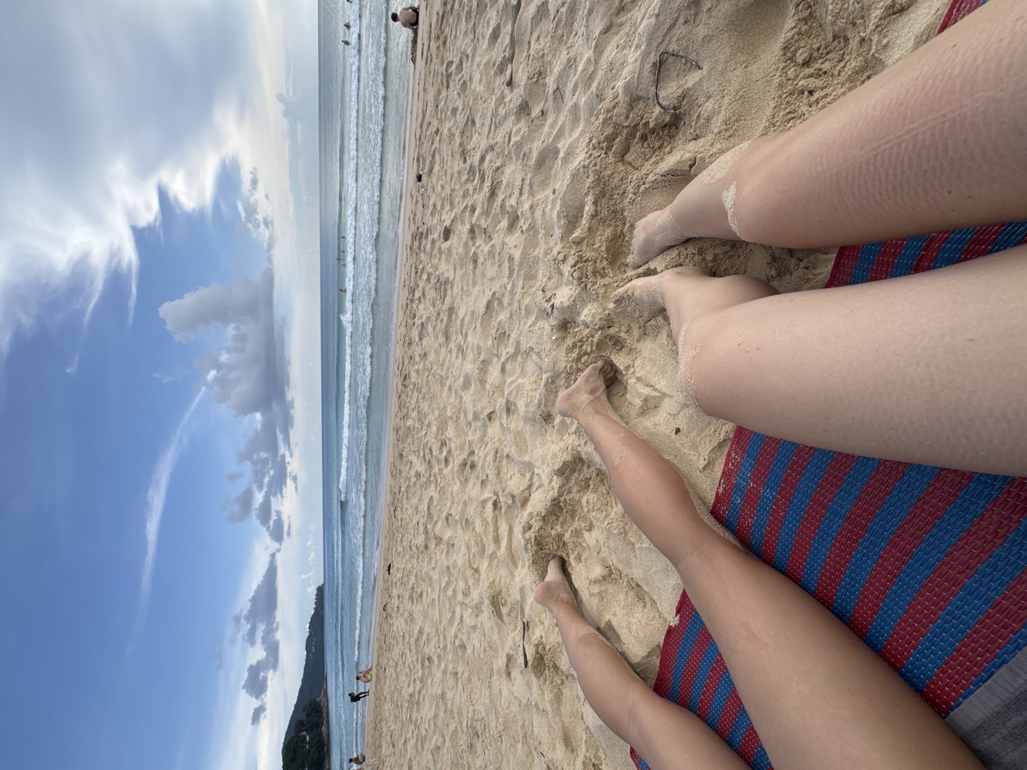 The image depicts a low-angle view of two individuals' legs resting on a sandy beach. A red and blue striped mat is positioned beneath their legs. The sand is lightly scattered on their skin. In the background, the ocean meets the horizon under a cloudy sky. Waves are visible approaching the shore, and several people can be seen scattered on the beach and in the water, some appearing to be swimming or surfing. Lush green hills are visible on the left side of the frame. The scene is set at Bang Tao Beach, in the Choeng Thale subdistrict of Phuket, Thailand.
