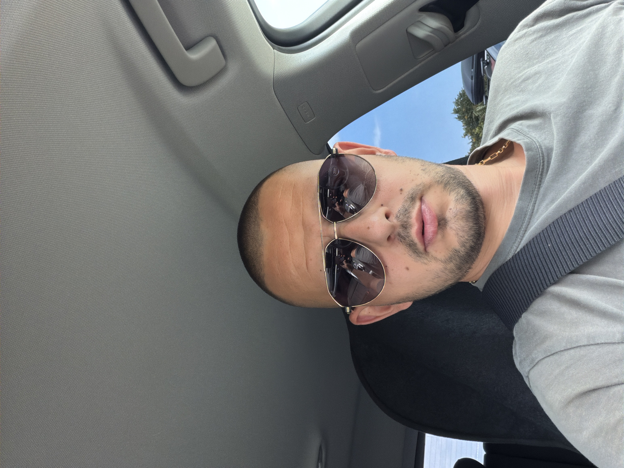 The image is a selfie taken from inside a car. A man with a buzz cut and sunglasses is in the driver's seat. He is wearing a grey t-shirt and a gold chain. The car's interior is visible, including the dashboard and a seatbelt. The sky and trees are visible through the car window. The location is the city of Burgas, Bulgaria.