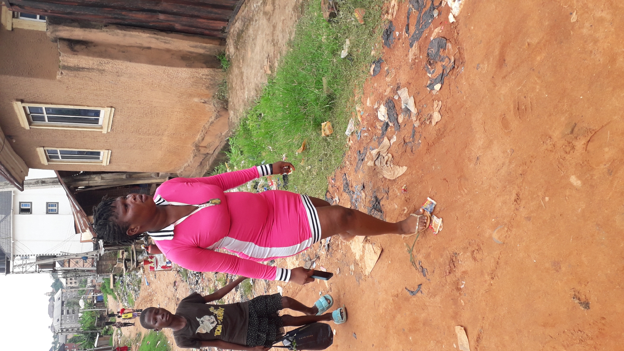 A woman in a pink dress with white and black stripes is standing on a dirt path in Nkpor, Nigeria. She is holding a smartphone in her right hand and is looking up. To her left is a young boy wearing a T-shirt with a cartoon character, shorts, and sandals. He is looking at the camera. The dirt path has scattered debris. The background features buildings, vegetation, and a view of the city.