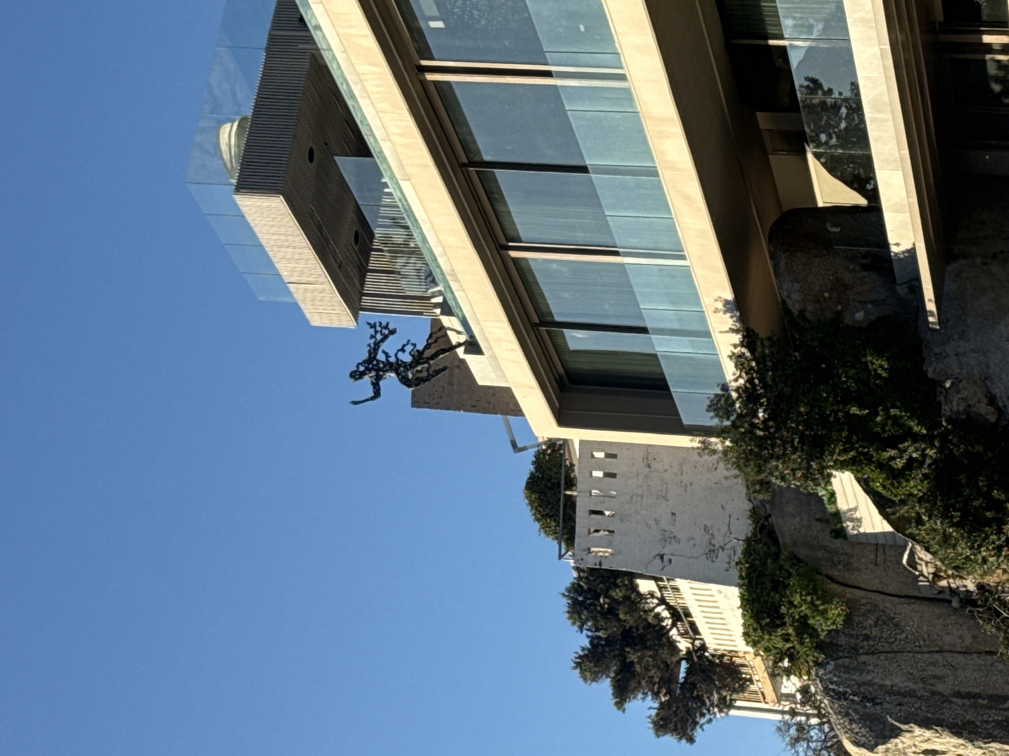 A low-angle shot showcases a modern building in what appears to be Cape Town, South Africa. The building features multiple glass-paneled levels with beige frames, juxtaposed against a clear blue sky. A metal sculpture of a figure is positioned near one of the upper levels. The building is set against a backdrop of natural elements including rocky terrain and lush green foliage. A section of a white building with numerous small square windows is visible beneath the modern structure.