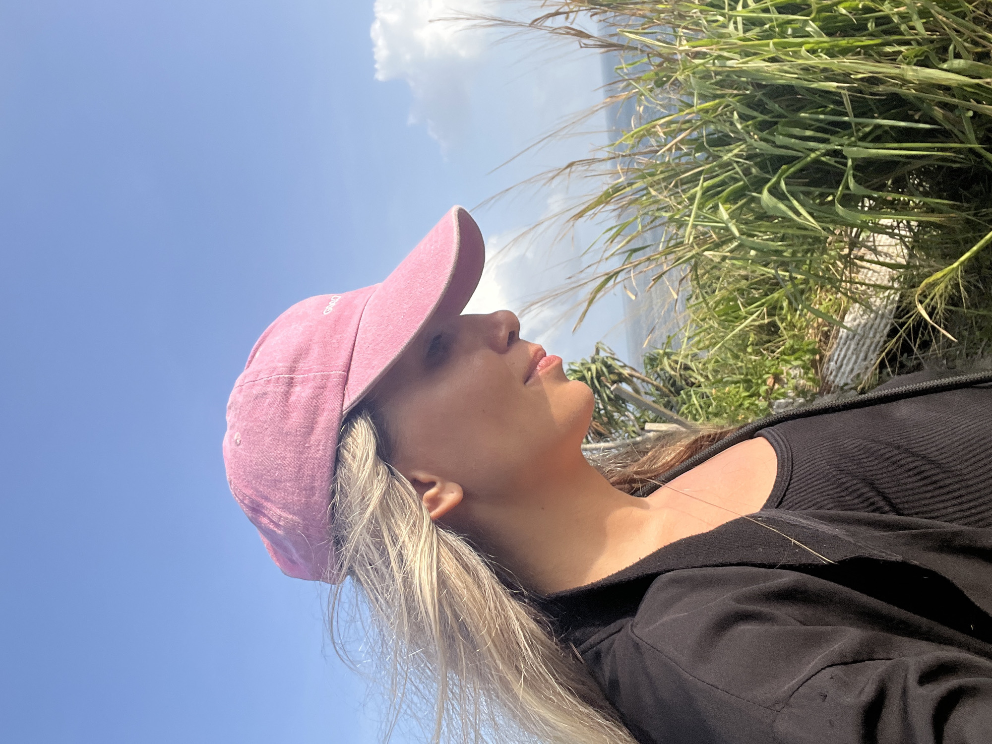 A woman wearing a pink baseball cap and a black jacket over a black ribbed tank top is pictured in profile, looking down and to the right. Her blonde hair is blowing in the wind. She is standing outdoors, with tall green grass in the foreground and the ocean and a partly cloudy blue sky in the background. The scene appears to be located in Talalla, Sri Lanka.