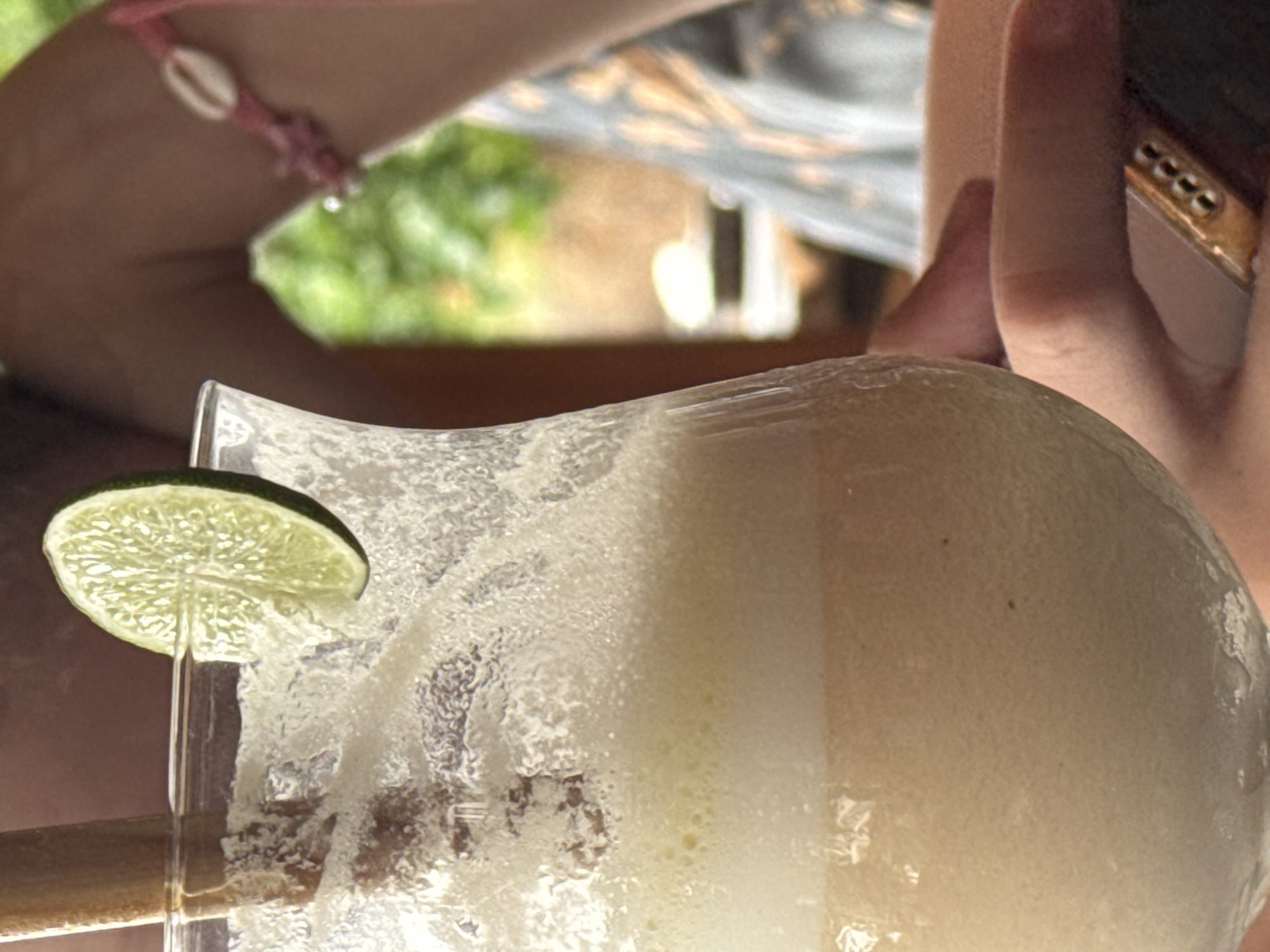 An extreme close-up shot focuses on a frothy, light-colored beverage in a clear, curved glass, garnished with a lime slice and a wooden straw. The drink is filled to the brim, with a thick layer of foam on top. In the background, out of focus, a person's arm is visible, adorned with a pink bracelet featuring a shell. The environment suggests an outdoor setting, possibly a tropical location, with lush green foliage visible. The overall impression is one of refreshment and relaxation, likely enjoyed in a warm climate. The image does not contain information about the city of Abang, Indonesia.