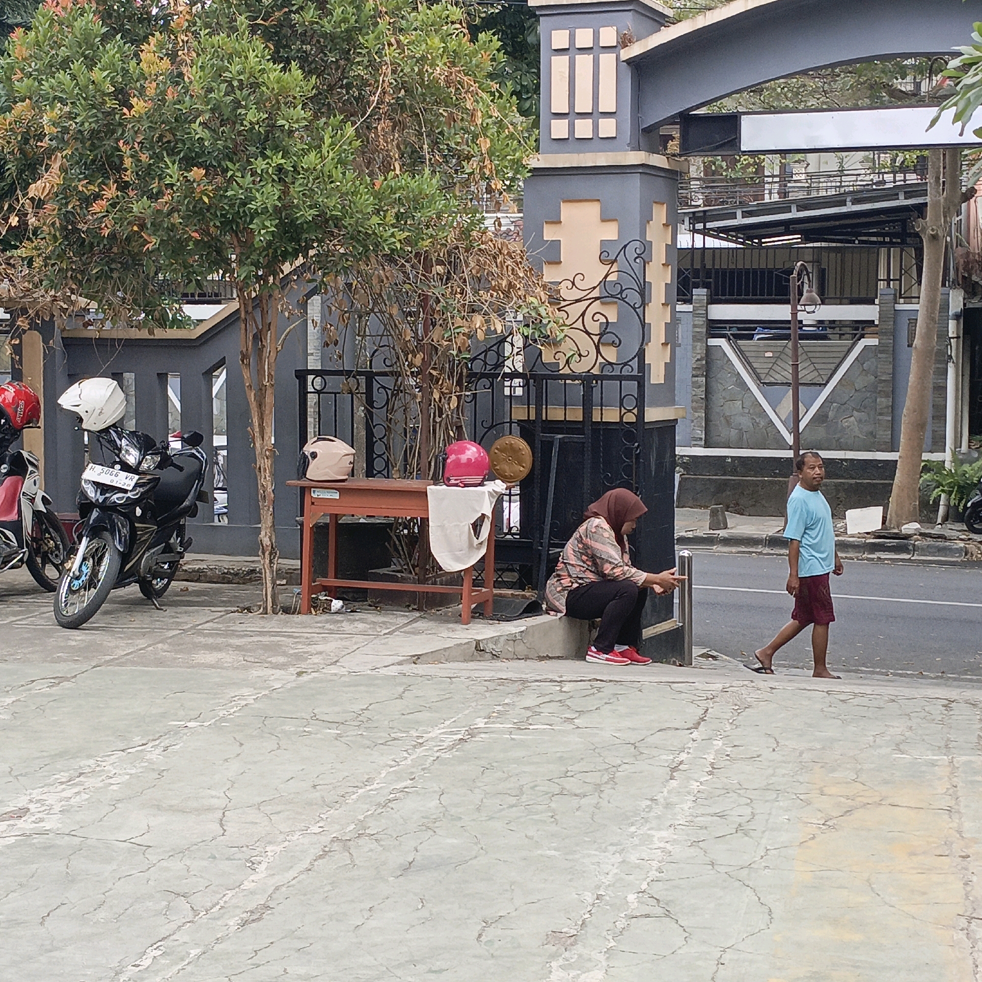 The image depicts a street scene in Salatiga, Indonesia, in front of a gated property with a prominent archway. In the foreground, a cracked concrete sidewalk is visible. To the left, two motorcycles are parked; one is black with a white helmet and the other is red and white with a red helmet. A woman wearing a brown hijab and a patterned shirt and black pants is crouched near a low, orange table, holding a phone. On the table are two helmets, one pink and one beige, and a circular, textured object. To the right of the woman, a young boy in a light blue t-shirt and dark red shorts walks on the sidewalk towards the street. Further to the right, a man in a light blue t-shirt and dark red shorts walks on the street. The background shows a gray gate with decorative beige accents, a tree with green and dry leaves, and buildings.