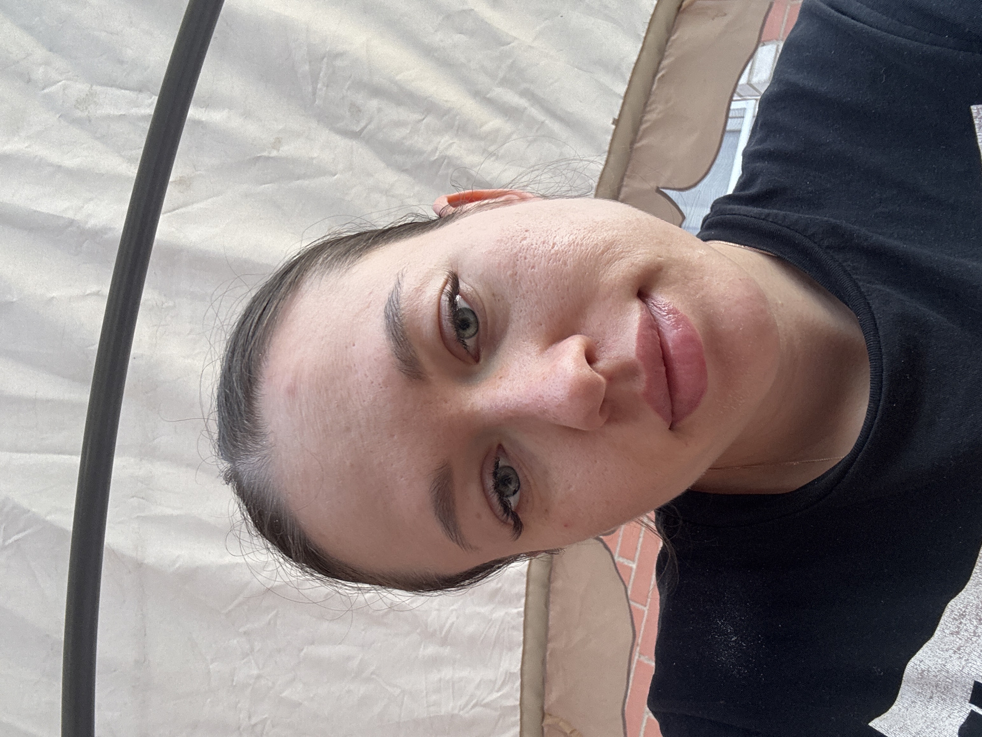 A selfie of a woman with light skin, gray eyes, and dark hair pulled back is angled towards the camera. She is wearing a black shirt. The background includes a beige tent-like material and part of a brick wall. The image was taken in Ферма, Russia.