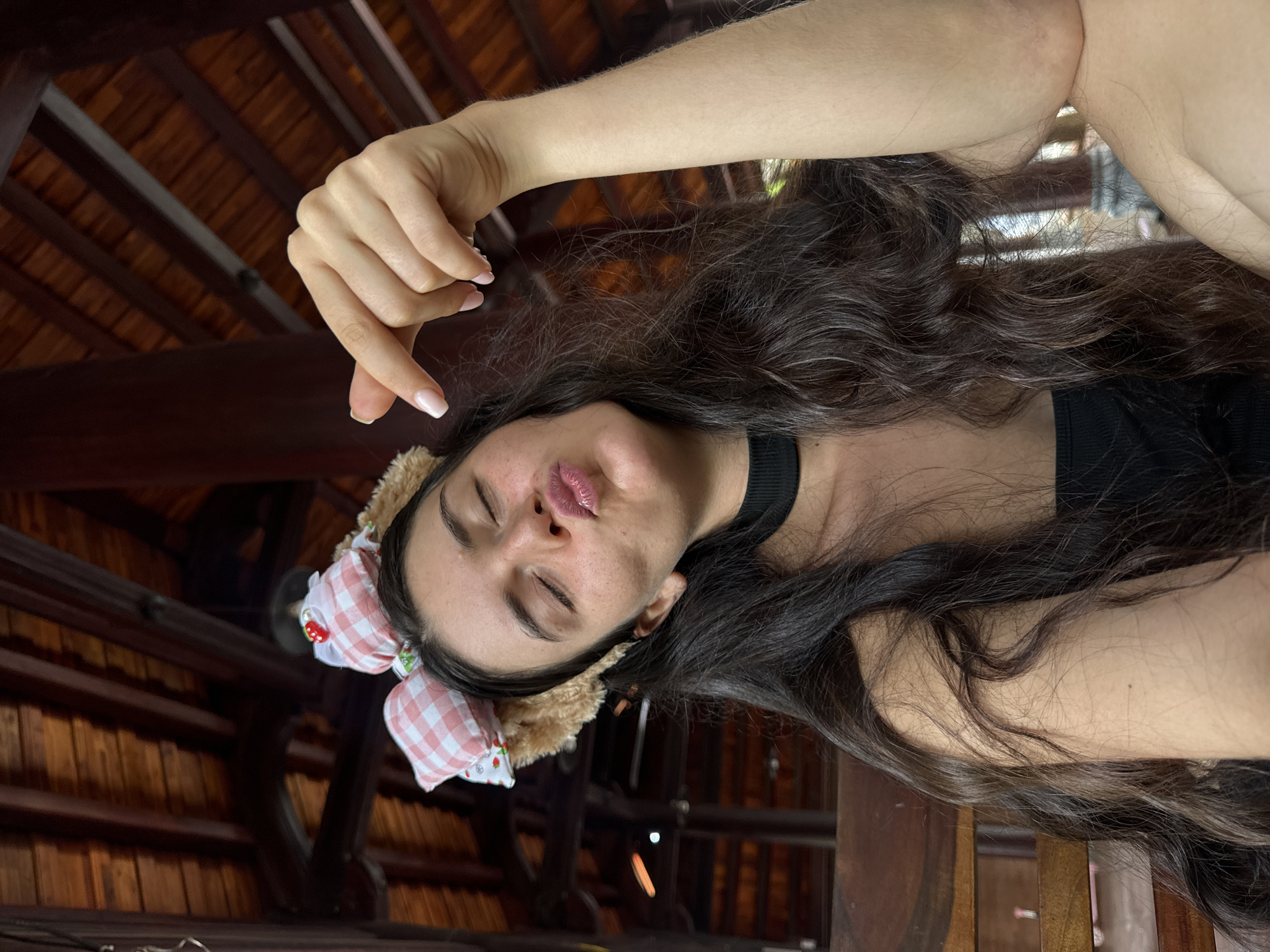 A young woman with long, dark, wavy hair and a black choker is posing for a selfie. She is wearing a headband with fluffy bear ears and pink and white checkered bows adorned with strawberries. Her eyes are closed, and her lips are pursed in a kiss. Her right hand is held up, with her index finger and thumb forming a small circle. She is sitting on a wooden bench in an outdoor setting with a wooden ceiling structure visible in the background. The lighting suggests it is daytime. The environment appears to be a shaded, covered outdoor area, possibly a restaurant or cafe, consistent with locations found in Mũi Né, Vietnam, which is known for its beaches and resorts.