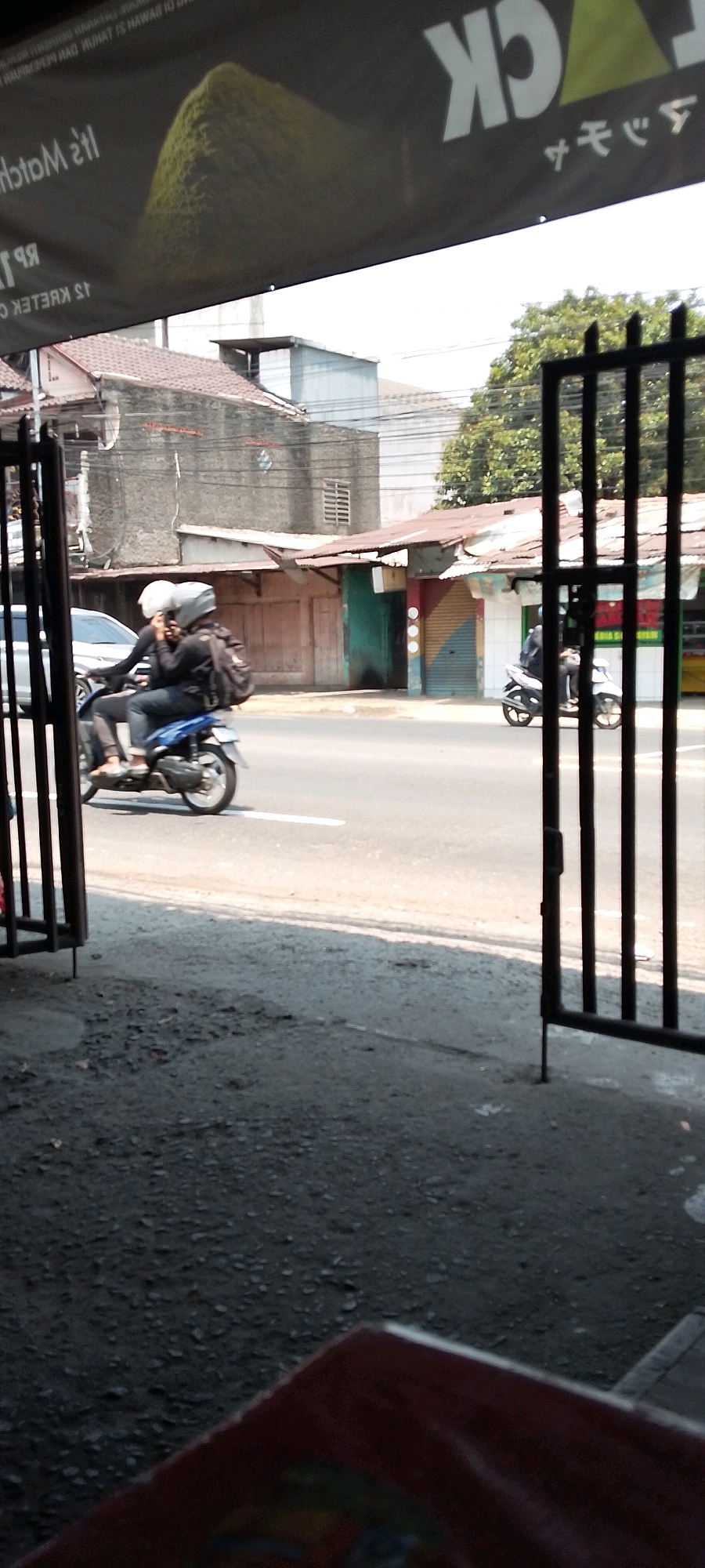 In Depok, Indonesia, two individuals on a blue motorcycle are captured from the side on a paved road. The driver, wearing a black outfit and helmet, is in front, while a passenger, also in black with a white helmet, sits behind. The motorcycle has a black backpack attached to its rear. The background reveals buildings with corrugated metal roofs, some appearing aged, and trees. In the foreground, a dark metal gate and rough ground are visible. A banner with text in English and Japanese is partially seen at the top, suggesting a commercial or advertising context. The scene takes place on a street with clear daylight, indicating it's daytime.