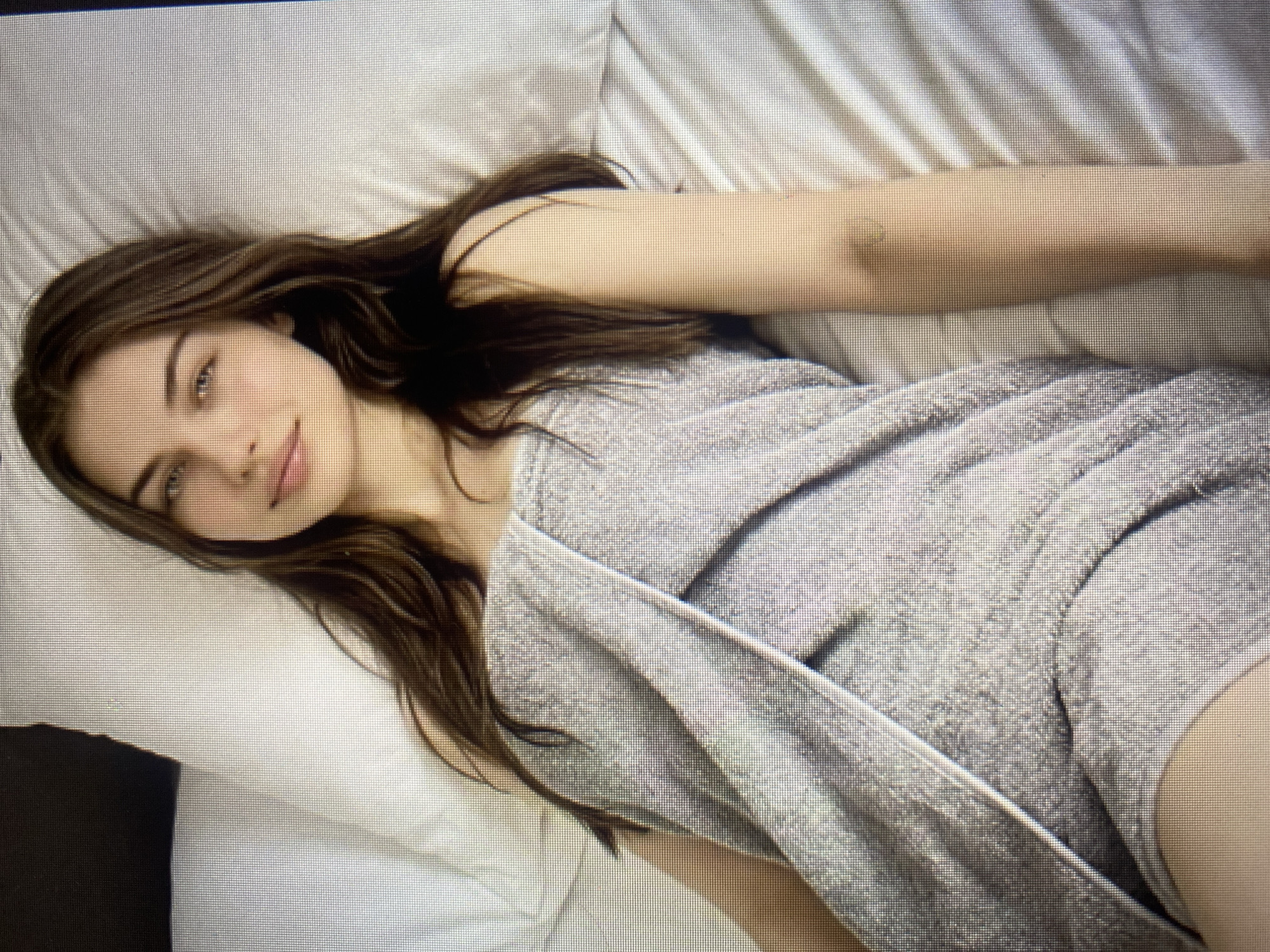 A young woman with long, brown hair and blue eyes is lying on a white bed, wrapped in a grey towel. Her head is tilted slightly to her left, and her gaze is directed towards the viewer. Her expression is relaxed and inviting. The room appears to be a bedroom, with white sheets and pillows on the bed. The lighting is soft and natural, creating a peaceful atmosphere. The scene is captured from a slightly elevated angle, providing a clear view of the woman and her surroundings. There are no other individuals or objects of note in the image.