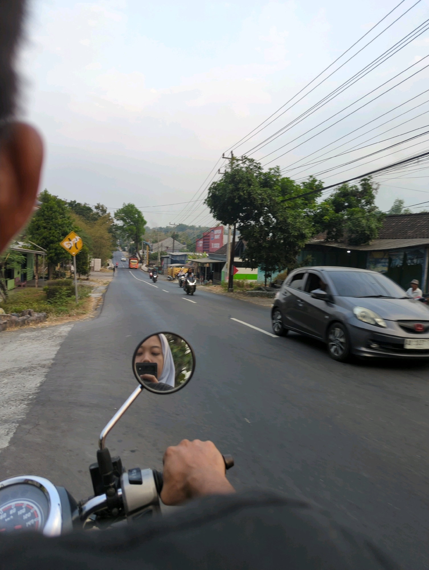 A POV shot from a motorcycle on a paved road, likely in Senggrong, Indonesia, shows a woman in a hijab reflected in the side mirror, holding a smartphone. The woman is looking into the mirror, and the phone is positioned to take a selfie. The motorcycle's handlebars and part of the dashboard are visible in the foreground. The road is lined with trees and buildings. Other motorcycles and a dark gray car are also on the road. The sky is overcast with power lines stretching across it. A yellow diamond-shaped traffic sign with black symbols is visible on the left side of the road. The scene appears to be during daylight.