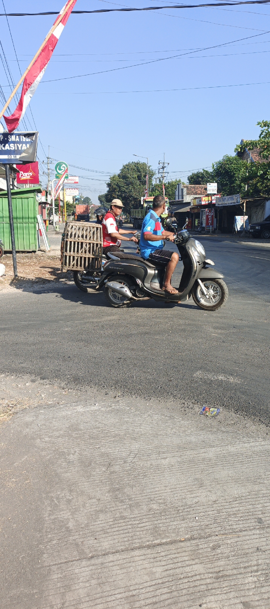 An outdoor, eye-level, full-shot photograph captures a bright, sunny day in Kasiyan, Indonesia, with a clear blue sky overhead. The scene depicts a street with asphalt paving and a concrete curb on the left. Two men are riding on a dark grey Honda Scoopy motorcycle. The man in the front, wearing a blue shirt and black shorts, is operating the motorcycle. The man behind him, wearing a red and white striped shirt and a straw hat, is carrying a large, wooden birdcage on his lap. The motorcycle is moving from left to right. In the background, there are various buildings and trees. Signs in Indonesian are visible, including one that reads "P. SMA ISLAM KASIYAN" and another with a green logo. There are also power lines and a utility pole. A green structure, possibly a kiosk, is on the left side of the road, and further down the road, a truck is visible. The overall impression is a snapshot of everyday life on a street in Kasiyan.