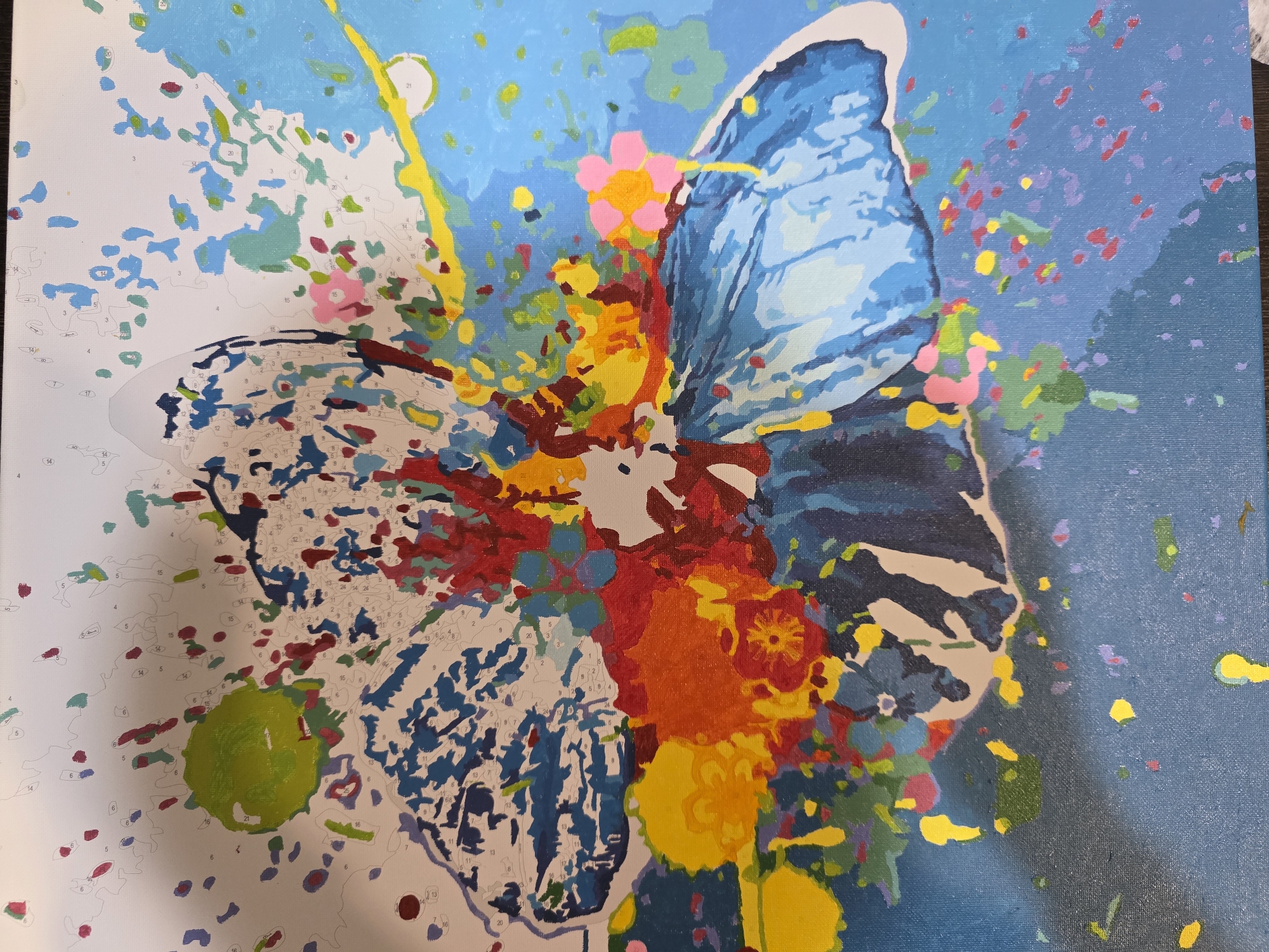The image depicts a partially completed paint-by-numbers canvas. The design features a butterfly with one wing painted in shades of blue and the other wing outlined with numbers and unpainted. Splashes of various colors surround the butterfly, some painted and some numbered. The background is partially filled with blue paint. There is no indication of location or connection to the city of Chaikovsky, Russia.