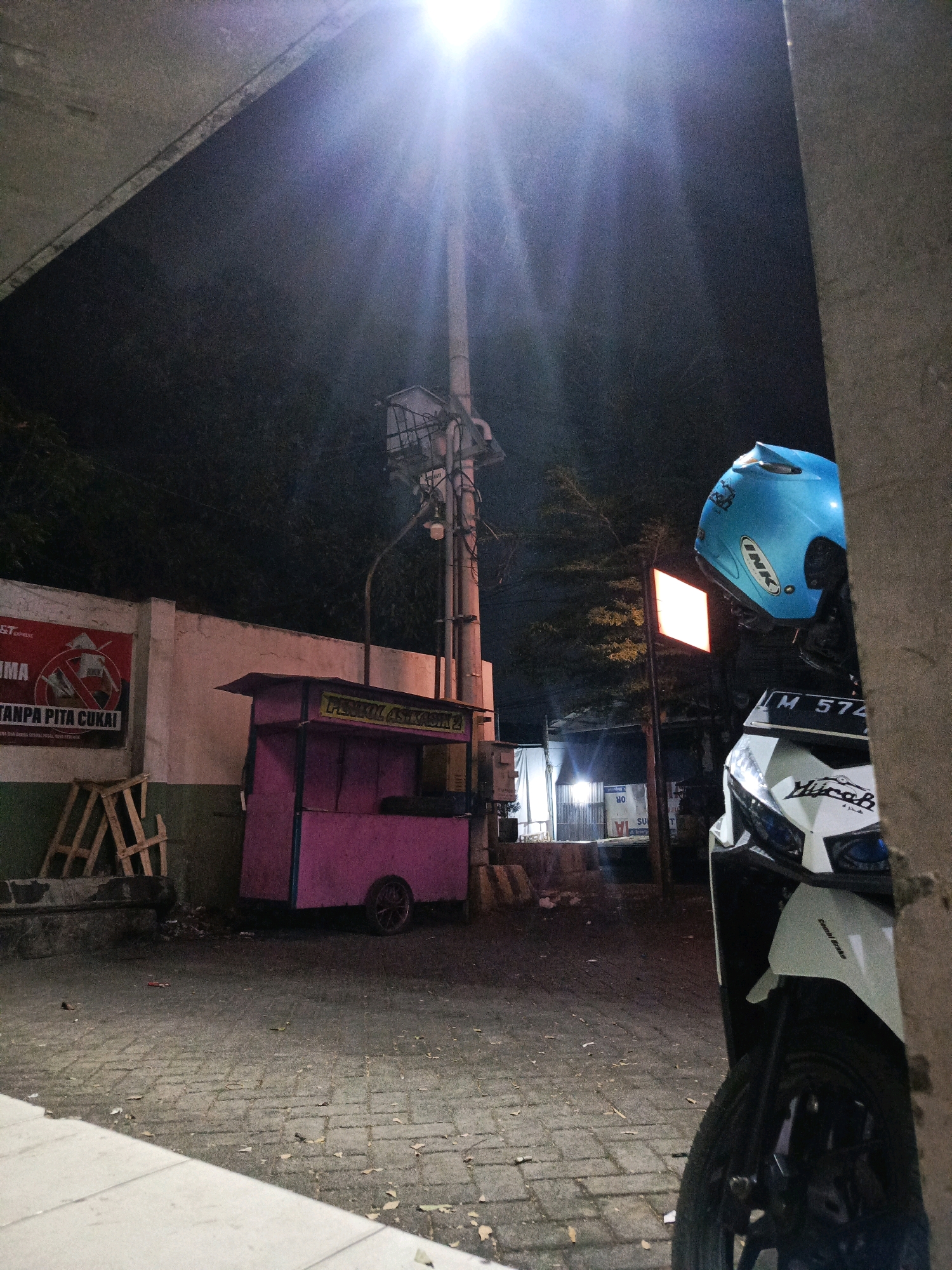 The image shows an outdoor scene at night in the city of Pamekasan, Indonesia, likely near a street or walkway. The foreground is dominated by the partial view of a white scooter with a blue helmet resting on its handlebars. The scooter has a license plate that reads "M 574". To the left, a pink food cart with a single wheel is visible, parked on a paved surface. Behind the cart, a concrete wall with a red and white poster is present. The poster features an image of a cigarette with a red cross over it and text that says "TANPA PITA CUKAI". A street light illuminates the scene from above, casting a bright glow. In the background, there are some trees and buildings, suggesting a moderately urban environment. The overall atmosphere is one of nighttime activity, possibly a roadside food stall or a parked vehicle.