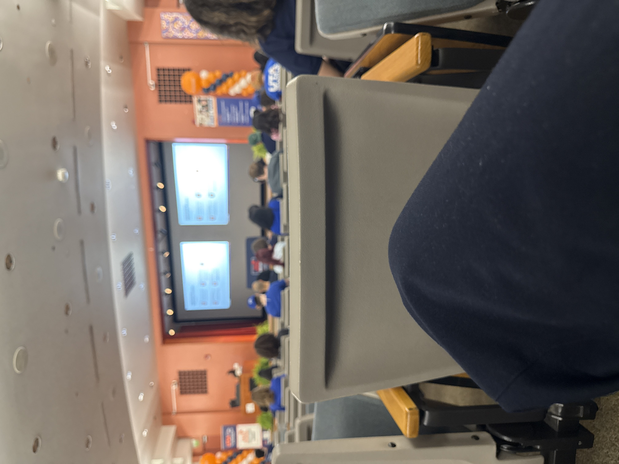 The image shows an auditorium in San Antonio, Texas, likely at the University of Texas at San Antonio (UTSA) based on visible school colors and logos. A presentation is displayed on two screens at the front of the room. People are seated in rows of chairs, facing the presentation. The vantage point is from one of the seats within the auditorium, focusing on the back of the seat in front and the presentation in the distance.