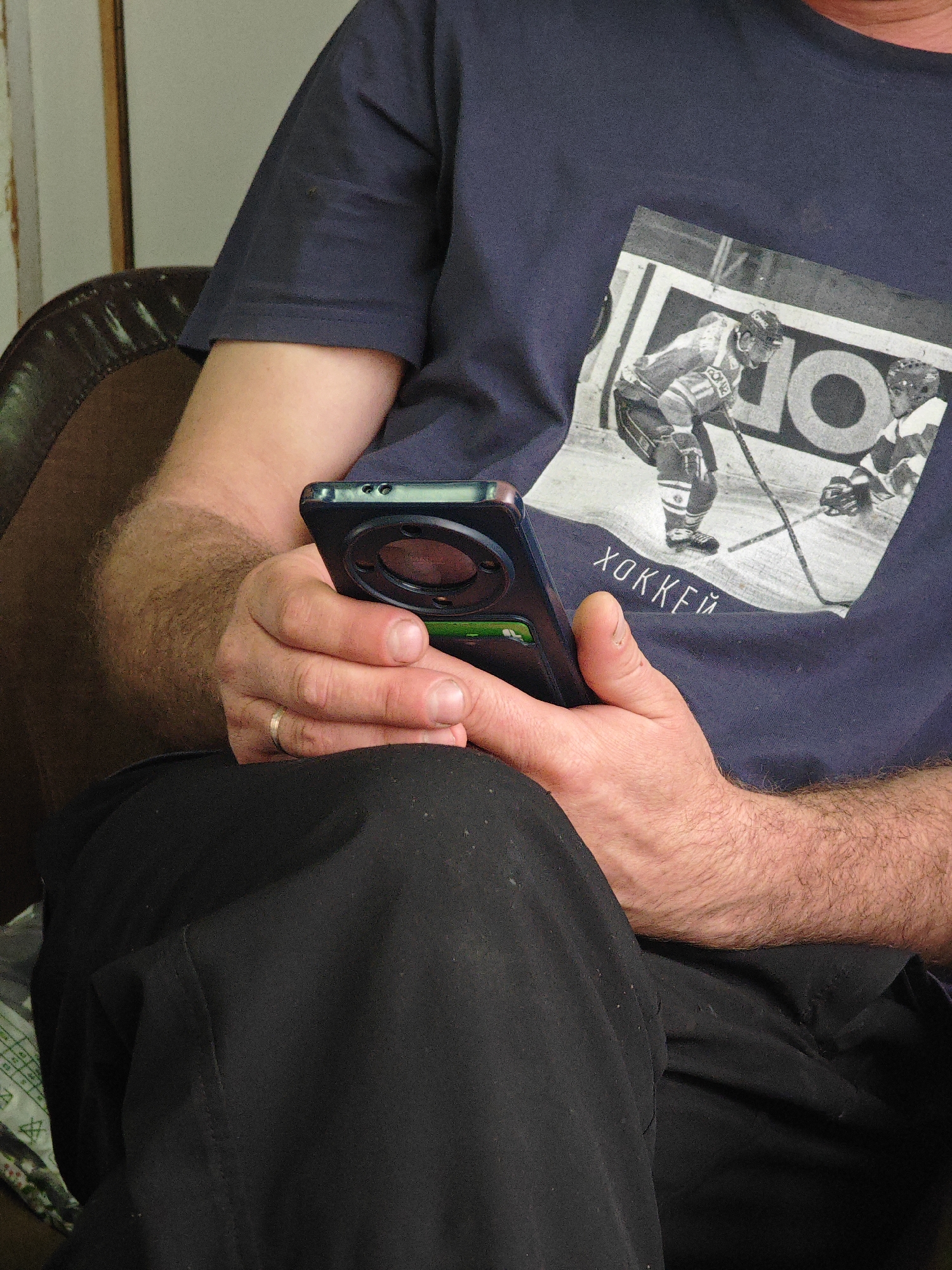 The image is a close-up shot of a person's torso and hands. The person is wearing a dark blue t-shirt with a graphic print of a hockey scene and the word "ХОККЕЙ" (Hockey in Russian). They are holding a black smartphone in their hands. The person appears to be sitting, as indicated by the dark fabric that could be clothing or upholstery. The background is blurred and indistinct, suggesting an indoor or shaded outdoor setting. There are no visible landmarks or environmental cues that would allow for identification of the location within Krasnoyarsk, Russia. The lighting is natural and appears to be from an overhead source. The image focuses on the interaction between the person and their phone.