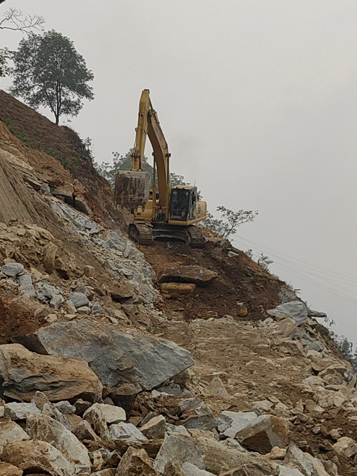 A yellow tracked excavator is positioned on a steep, excavated dirt and rock slope in Trung Chải, Vietnam, indicating ongoing earthmoving or construction activity. The excavator's bucket is lowered, partially obscured by the terrain, and a plume of exhaust smoke rises from its engine area. The slope consists of exposed reddish-brown soil and numerous scattered boulders and fractured rock fragments, predominantly gray and brown, suggesting recent displacement. A path or cleared area of dirt and smaller rocks extends downwards and forwards from the excavator's position. On the upper left portion of the image, a solitary deciduous tree with green foliage stands atop the ridge. Sparse smaller trees and vegetation are visible elsewhere on the higher slopes. The sky is uniformly overcast and light gray. In the background on the right, parallel power lines are faintly visible against the sky. No individuals are present in the frame.