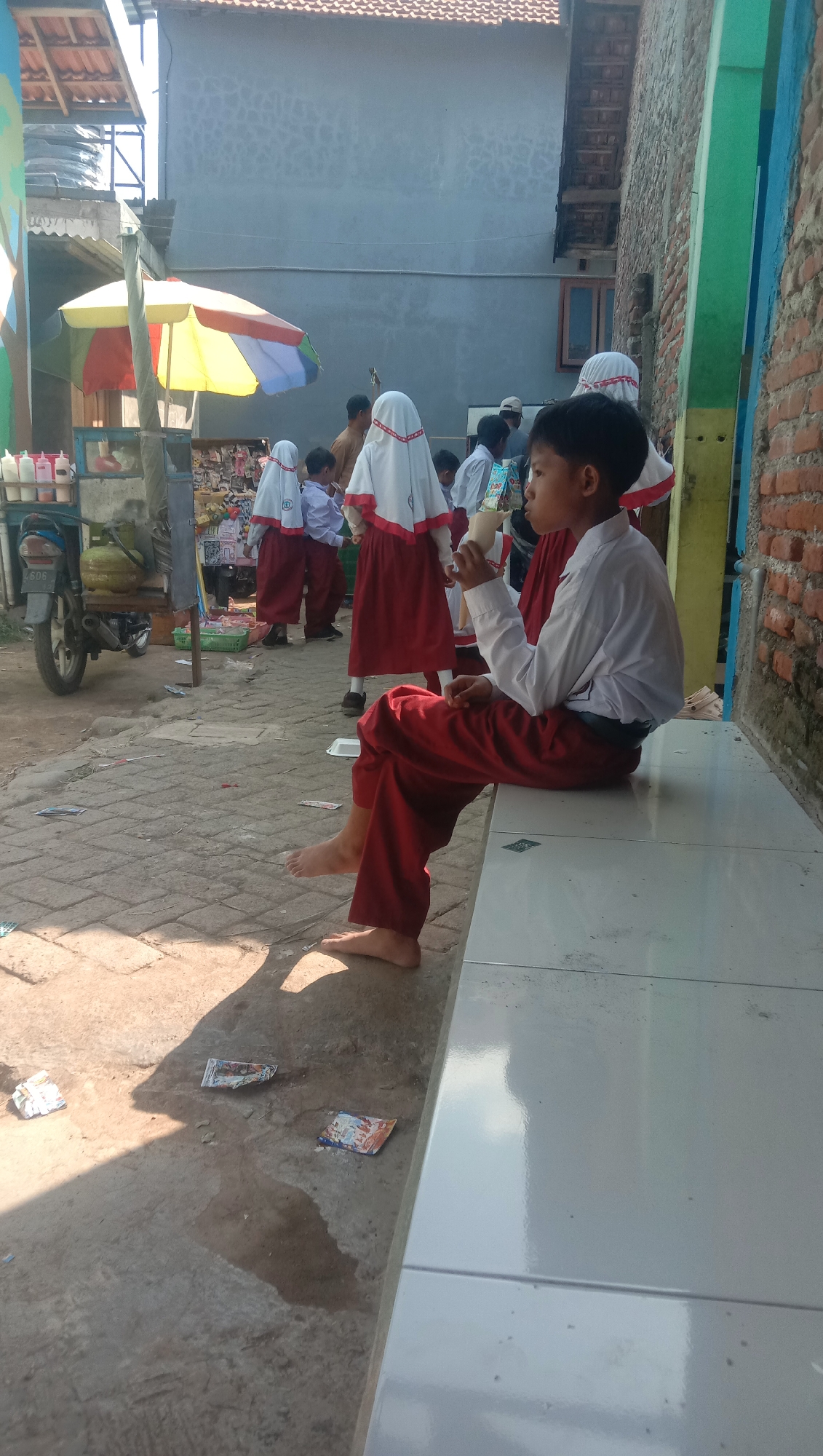 The image depicts a street scene in Karang Anyar, Indonesia, on a sunny day. In the foreground, a young boy wearing a white shirt and red pants is sitting on a white tiled bench, holding a light-colored object, possibly a food item or drink, to his mouth. He is barefoot. To his left, the ground is paved with interlocking bricks, and several pieces of litter are scattered on it. A shadow of the boy falls across the ground.
In the background, several other children, presumably students, are visible. They are wearing white shirts with red accents and red pants or skirts, and some are wearing white headscarves. They appear to be gathered near a stall or kiosk under a colorful umbrella, which is yellow, red, and blue. Behind the children, a brick wall is prominent. To the far left, a motorcycle is parked. The overall environment suggests a casual setting, possibly near a school or a marketplace.