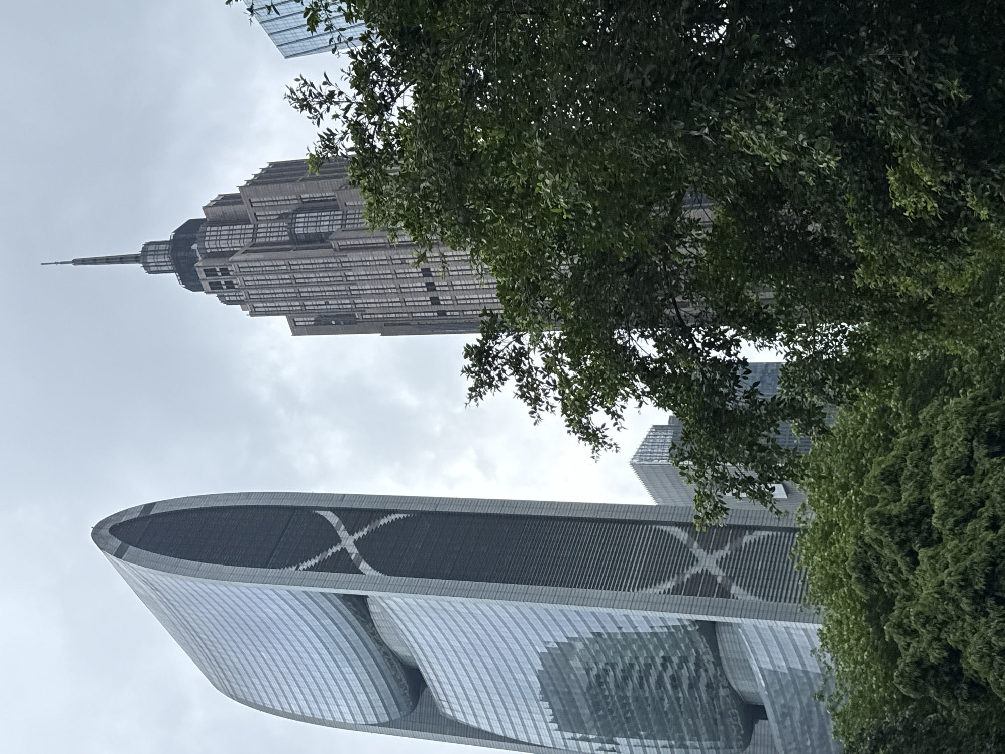 The image shows a view of modern skyscrapers in Tianhe District, China, on an overcast day. In the foreground, lush green trees obscure the lower portions of the buildings. To the left is a tall, curved skyscraper with a distinctive glass facade, featuring a large elliptical opening near the top, adorned with an "X" pattern. To the right of this building is a more traditional, stepped skyscraper with a spire, reminiscent of art deco architecture, its facade composed of numerous rectangular windows. Other, less distinct buildings can be seen in the background, also featuring glass exteriors. No people are visible in the frame. The sky is a uniform grey, indicating cloudy weather.
