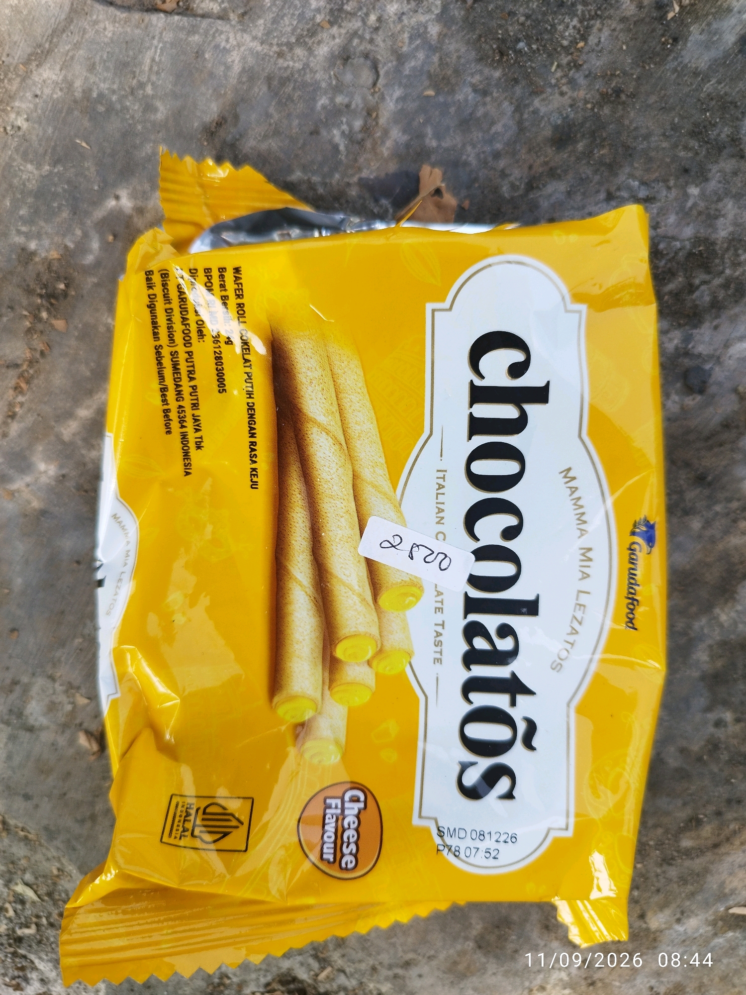 The image shows a yellow package of "Chocolatos" wafer rolls with a cheese flavor. The package is lying on a textured, gray surface that appears to be concrete or pavement. The lighting suggests it is outdoors during the day. There are some dried leaves scattered on the ground around the package. The brand logo "Garudafood" is visible on the right side of the package. Text on the package indicates it is a product of Indonesia, and a halal logo is present. A handwritten price tag reading "2800" is affixed to the front of the package. The timestamp "11/09/2026 08:44" is superimposed on the bottom right of the image, indicating a future date and time.