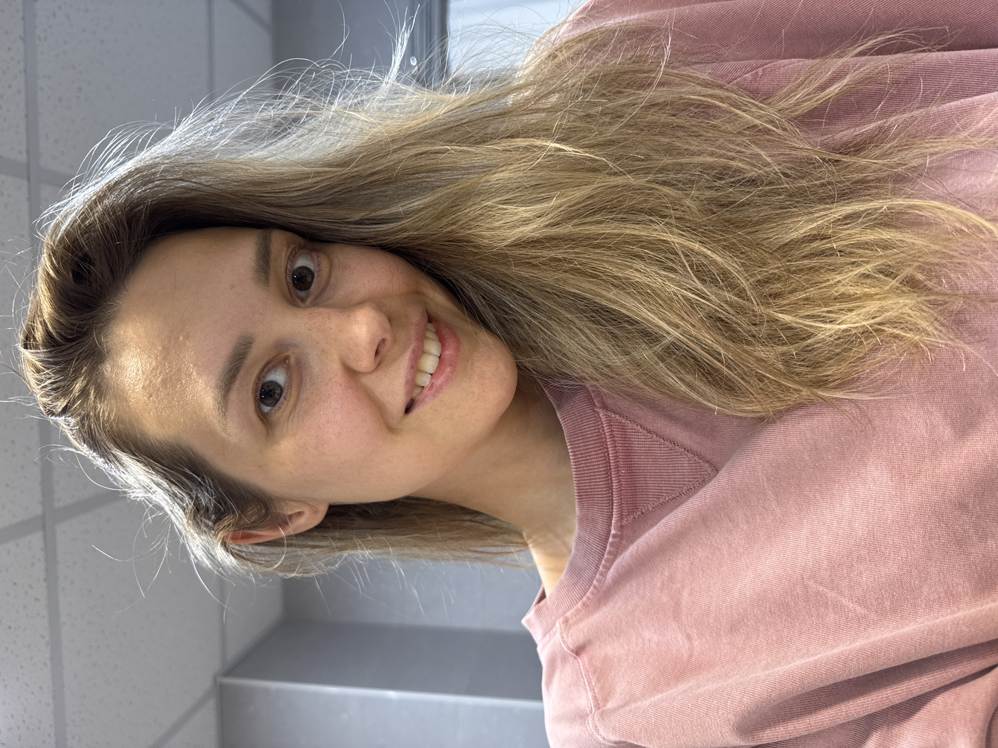 A young Caucasian woman with blonde hair and brown eyes smiles at the camera. She is wearing a pink crew neck sweatshirt. The background appears to be an indoor environment with a tiled ceiling and gray walls. The lighting suggests a bright, possibly artificial, light source. The image appears to be a selfie, taken from a slightly high angle. There is no information available about the specific location in Nebug, Russia.