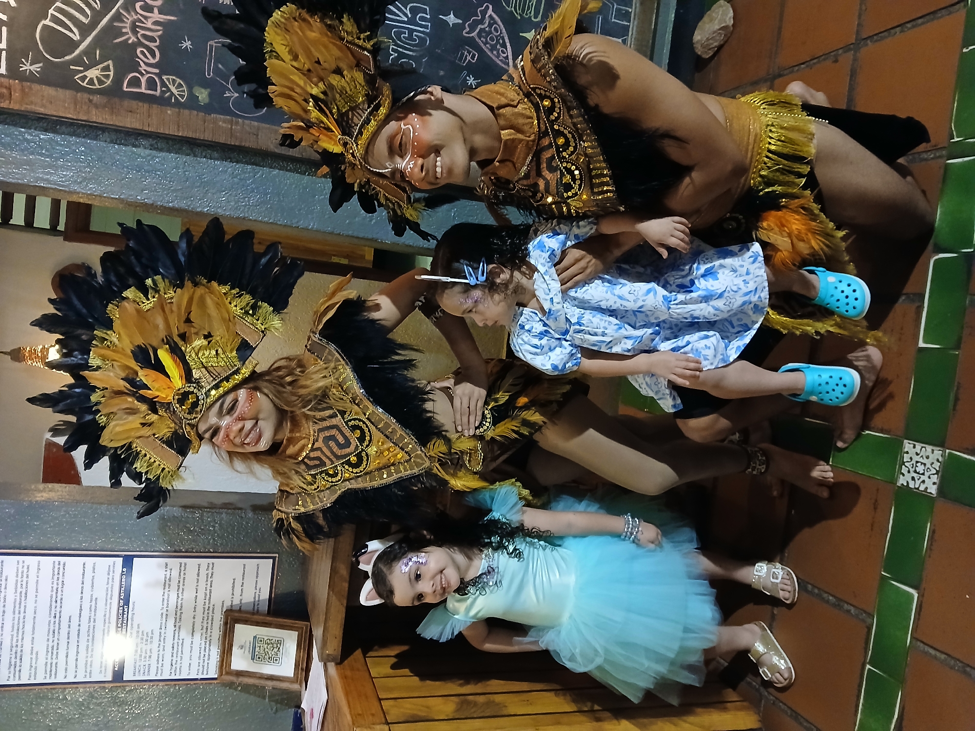 Here's a detailed description of the image:
**Scene:** The photo captures a group of people inside what appears to be a restaurant.
**People:**
* Two adults are dressed in elaborate carnival-like costumes featuring headdresses with feathers and gold detailing. They have painted faces.
* Two young girls are also present. One is dressed in a blue dress and the other in a blue tutu. Both have glitter on their faces.
**Objects:**
* The adults wear ornate headdresses with black and golden feathers and matching decorative collars.
* One girl is in a light blue dress. The other is in a sparkly blue tutu with white ears attached to her hair.
* A sign with the restaurant's policies is visible on the wall. A QR code is also displayed on the sign.
* Behind them is a chalkboard with "Breakfast" written on it.
* The floor is tiled in a red and green pattern.
**Actions and Interactions:**
* The adults are interacting with the children, possibly posing for the photo.
* The group appears to be celebrating or participating in a carnival event.
**Environment:**
* The location is indoors, likely in a restaurant, as indicated by the signs and the decor.
**Location Suggestion:**
* Given the description and the attire of the adults, it is possible the photo was taken in a restaurant in Porlamar, Venezuela, as suggested. Carnival is celebrated in this city.